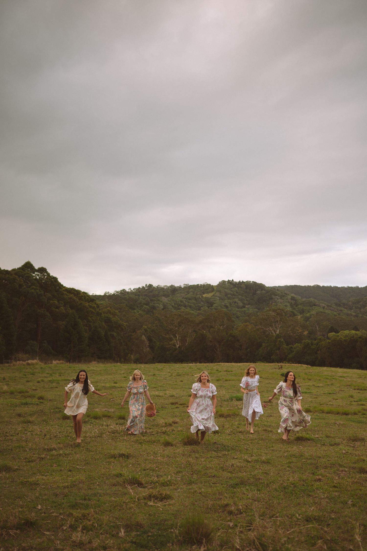 Five women in white floral dresses running through a green field with forested hills under cloudy skies.