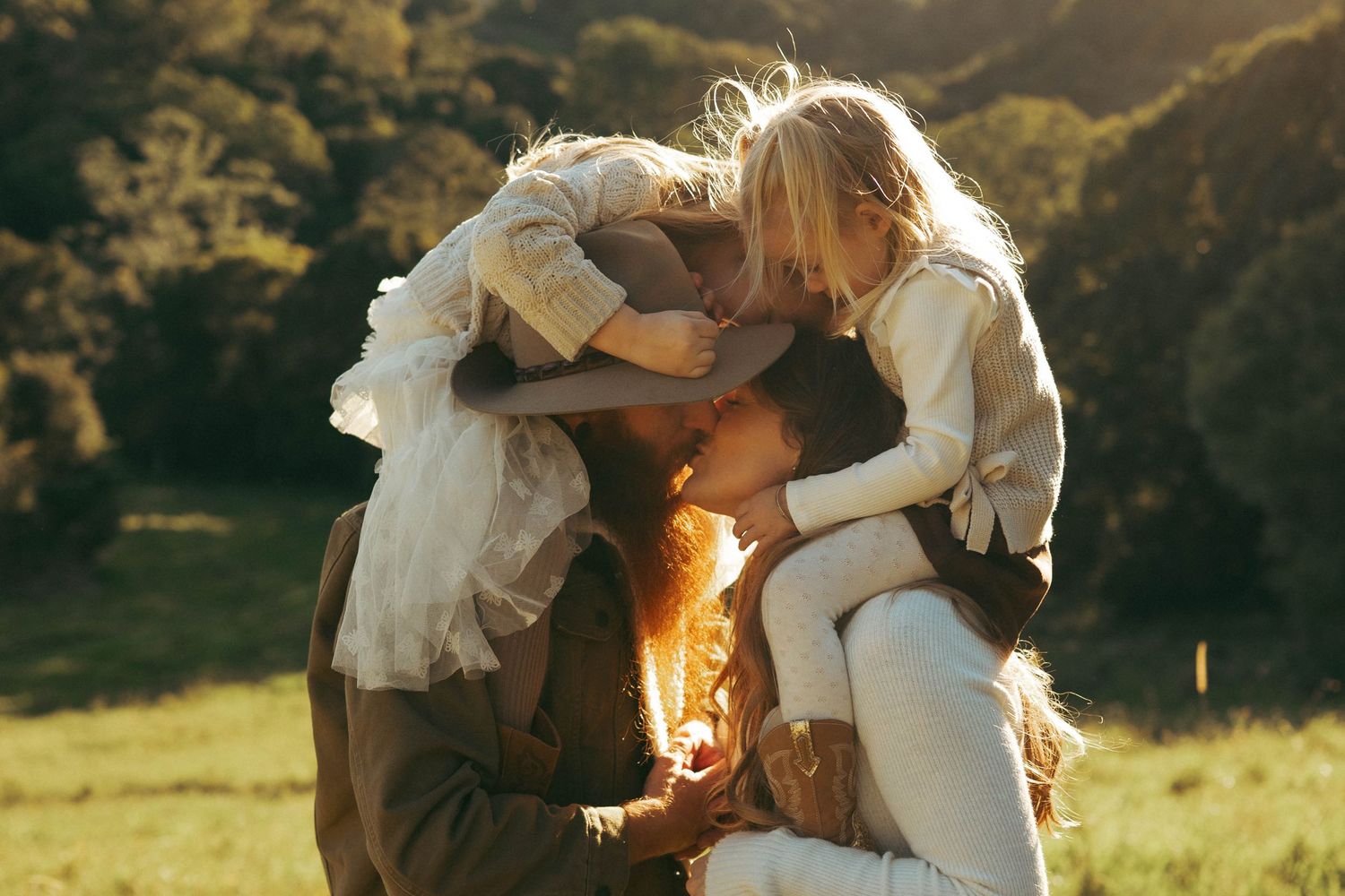 Family embracing outdoors in golden sunlight, two blonde children hugging parent in a lush green field.