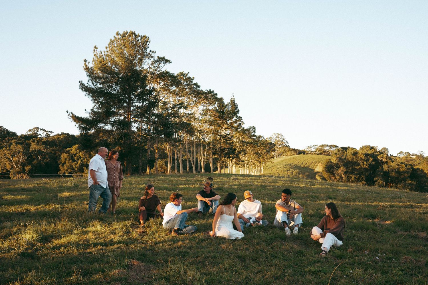 Group of people sitting together on a grassy field surrounded by tall trees during golden hour.