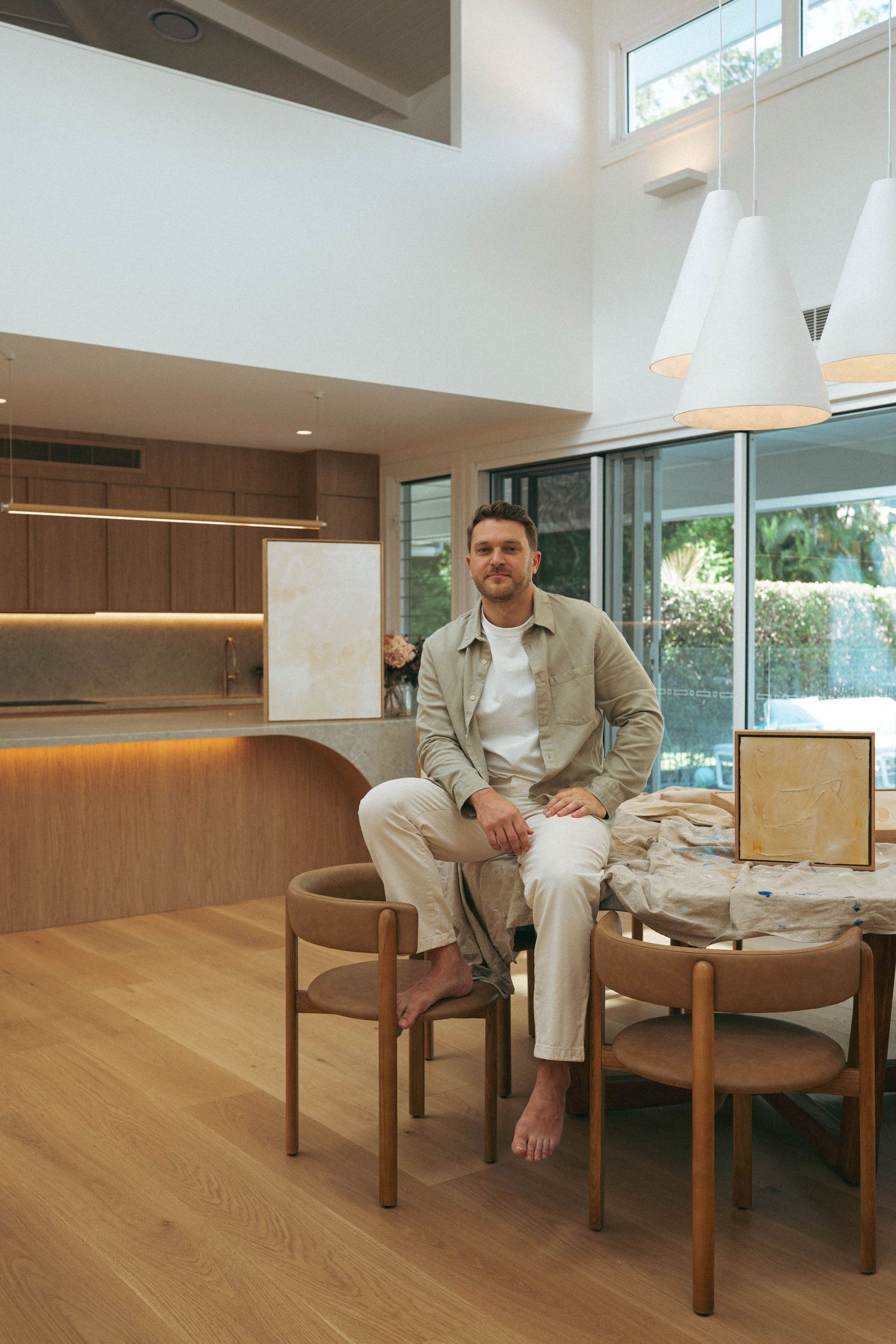 Man sitting barefoot at a wooden dining table in a modern open-plan home with high ceilings and warm timber finishes.