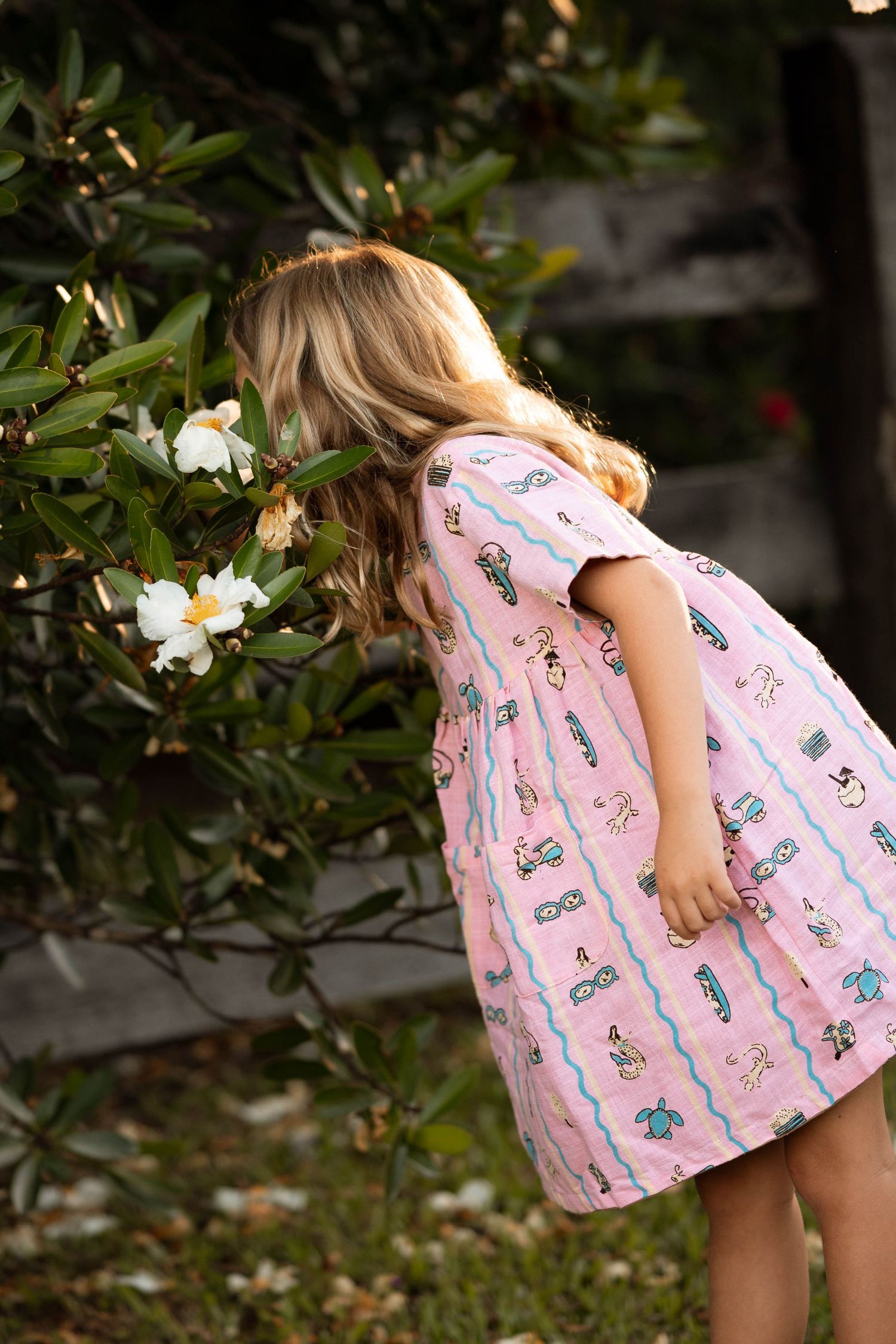 Young girl in pink printed dress smelling white flowers on a green bush outdoors.