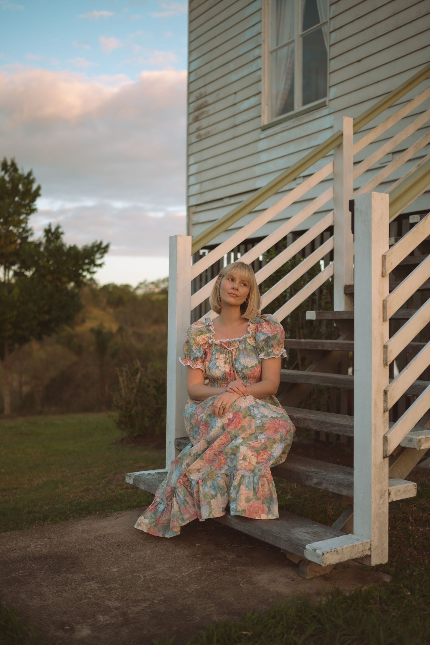 Woman in floral dress sitting on white wooden steps of a rustic building at golden hour.