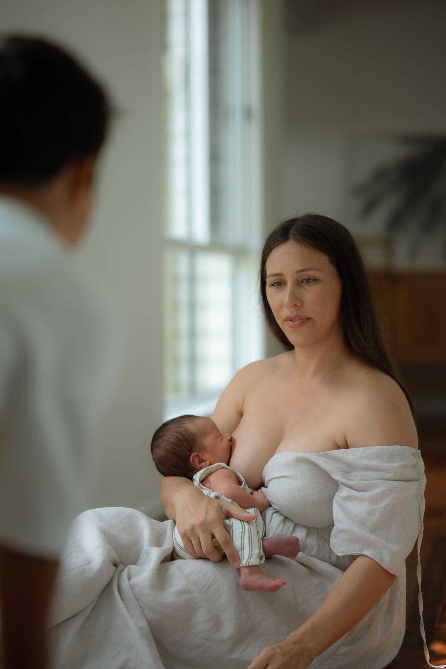 Mother in white off-shoulder top breastfeeding newborn baby while partner watches in bright room.