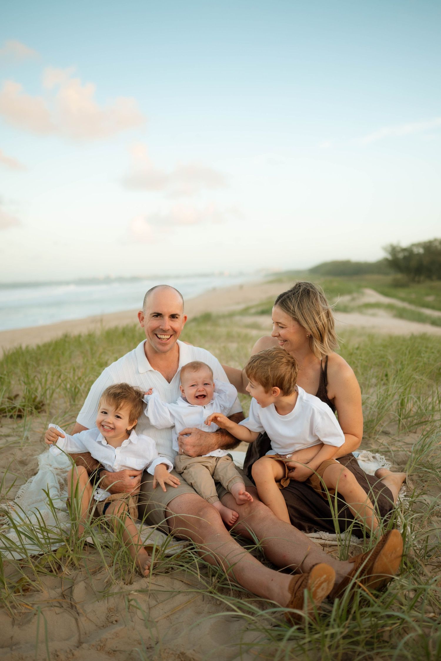 Happy family of five with three young boys laughing on beach sand dunes.