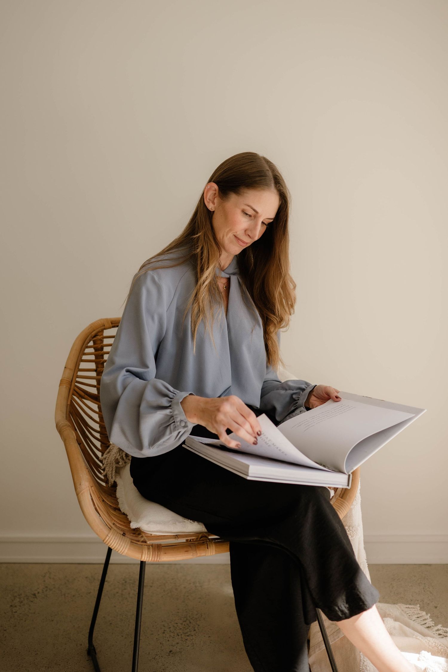 Smiling woman in grey blouse flipping through a large photo album while seated in a wicker chair.