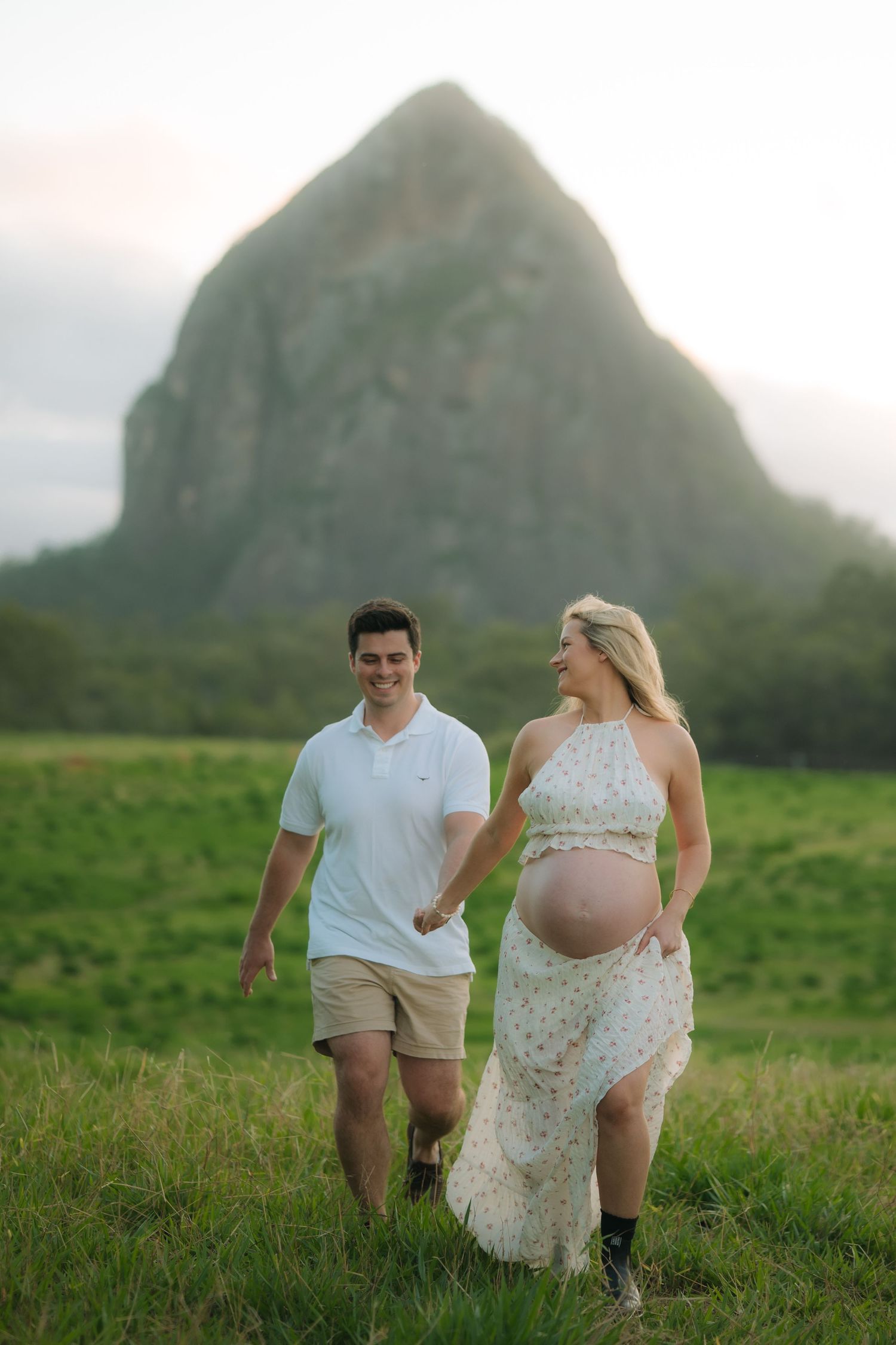 Happy couple walks hand in hand through lush green field with large rock formation visible in the background.