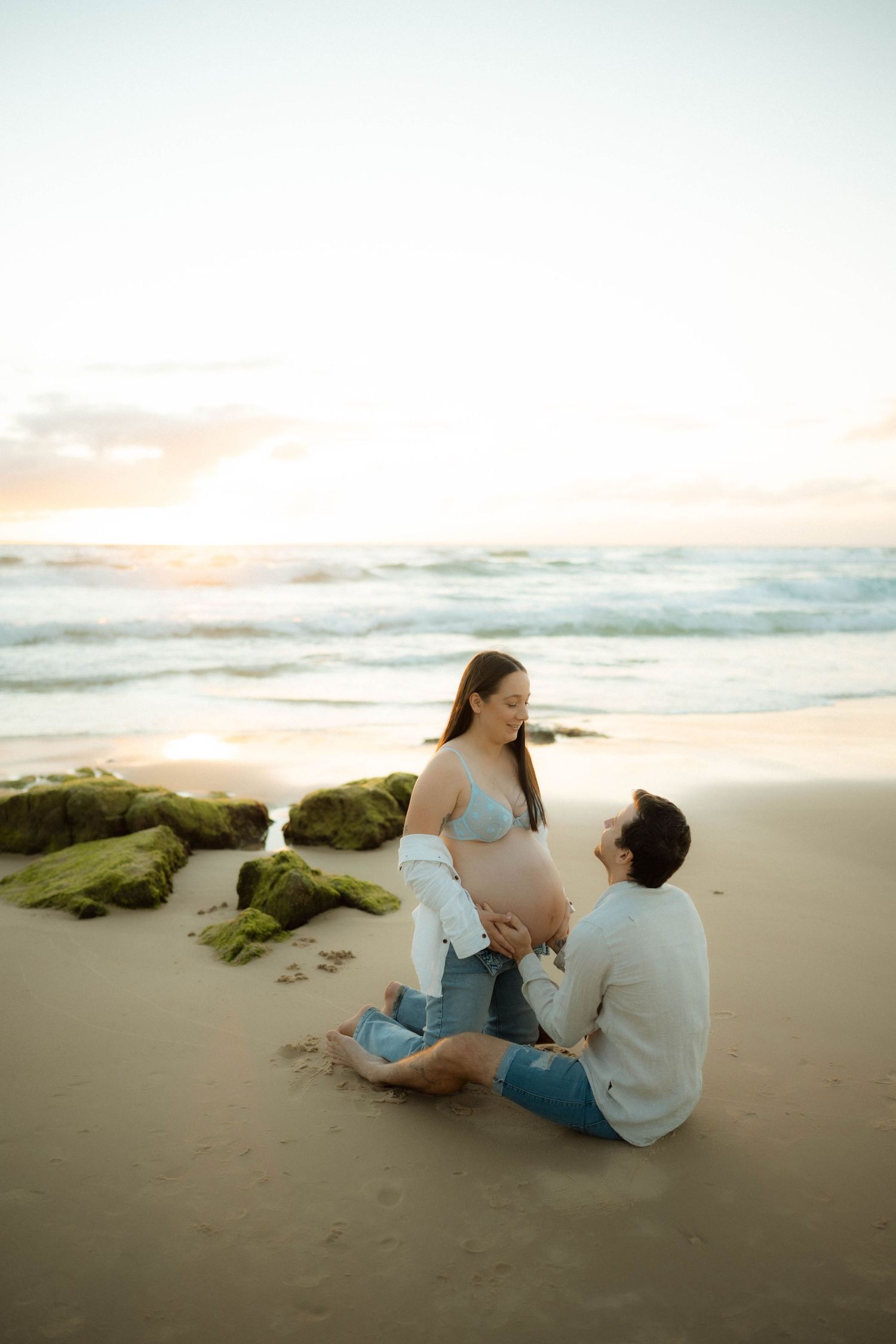 Man sitting on beach gazing up at standing pregnant partner during golden hour sunset.
