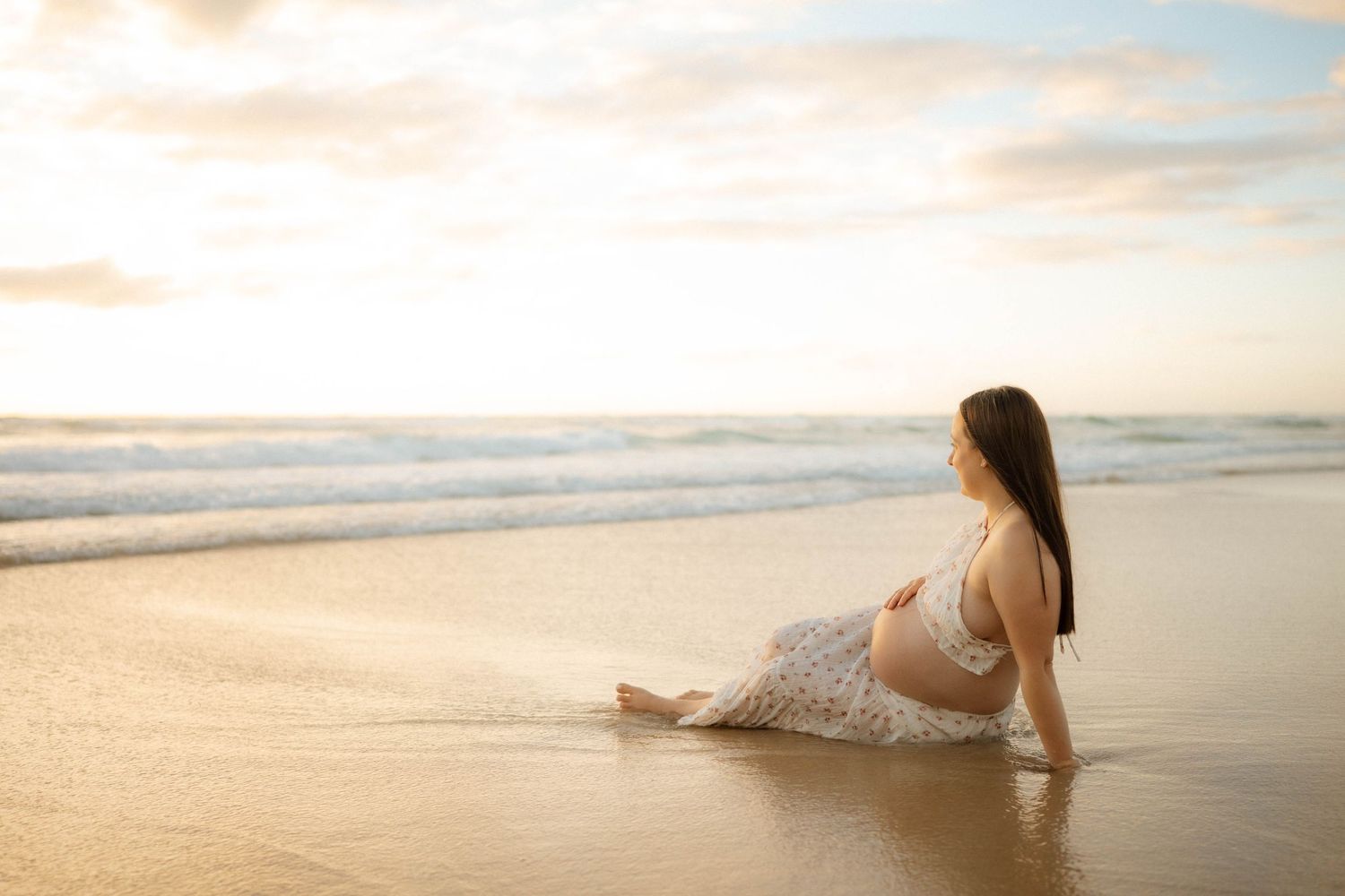 Pregnant woman in floral dress relaxing on sandy beach at dusk, cradling baby bump near ocean.