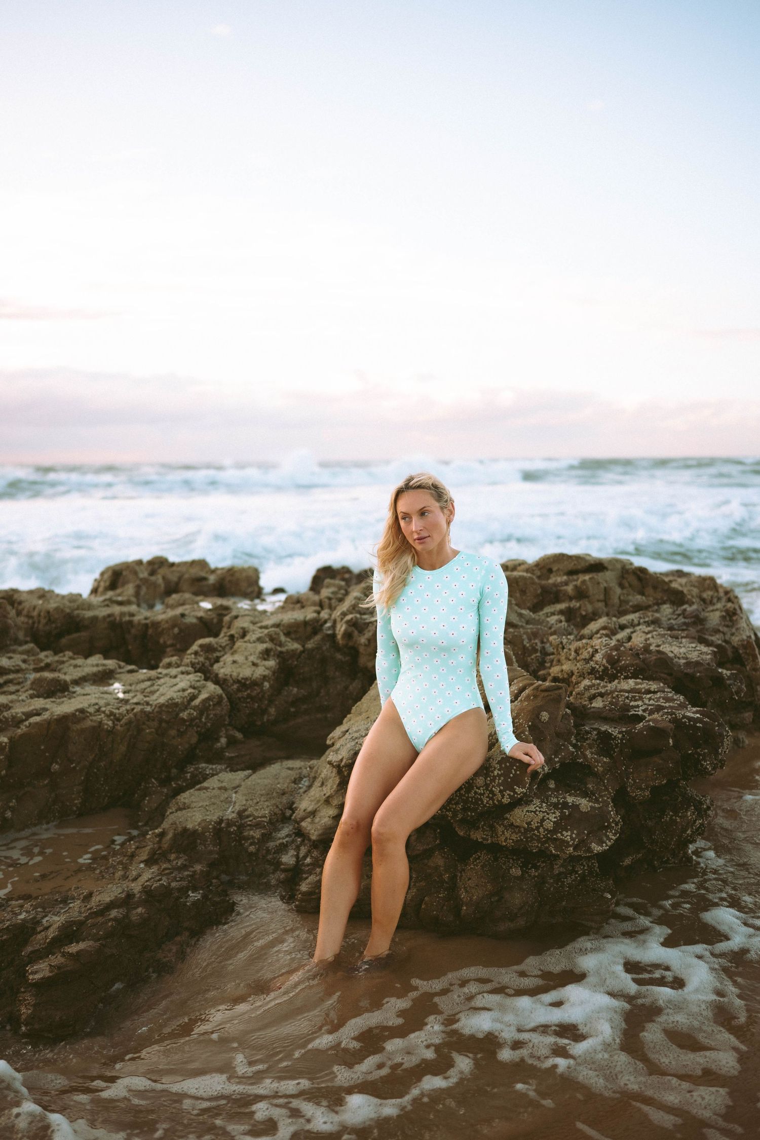 Blonde woman in light blue long-sleeve one-piece swimsuit leaning on coastal rocks at beach.