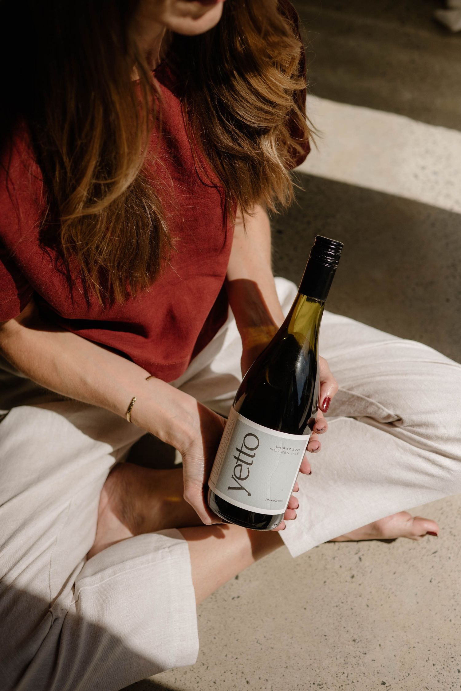 Woman in red top holding a bottle of Yetto wine while sitting cross-legged on the floor.