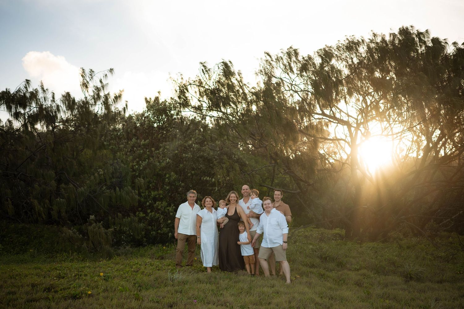Family group of seven standing in a grassy field at golden sunset with lush green trees in background.