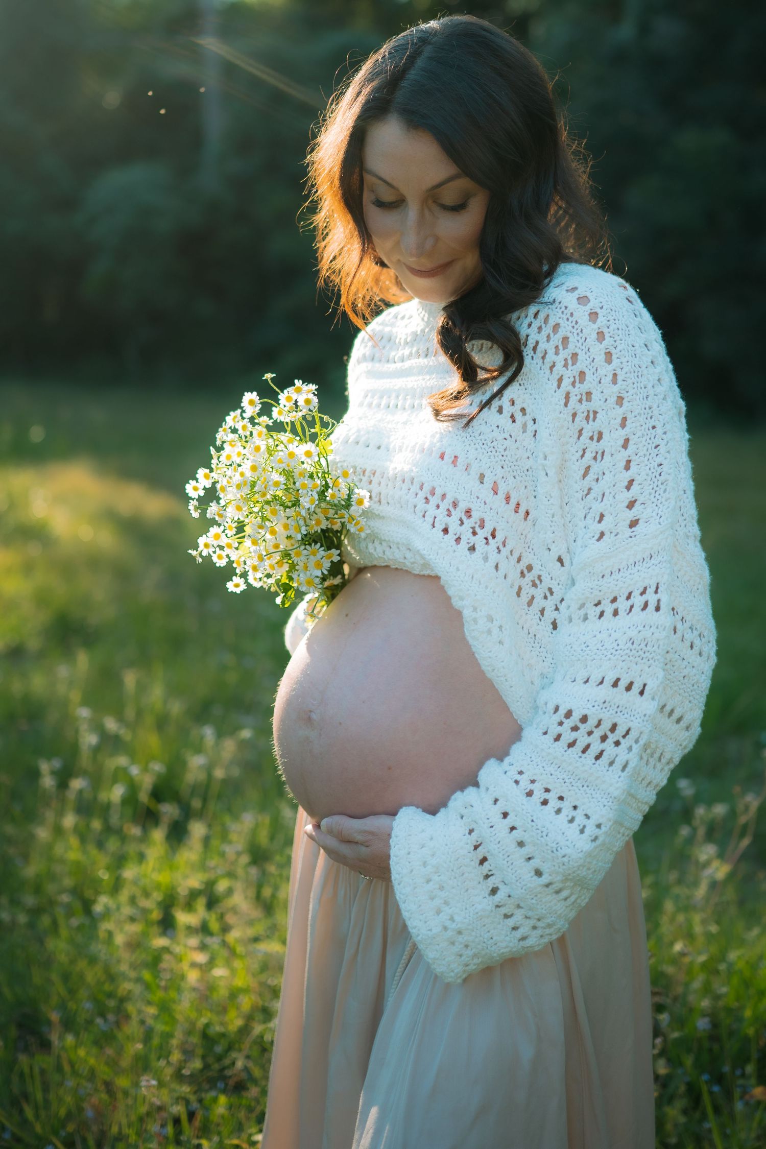 Pregnant woman in white knit sweater cradling her bump and holding a bouquet of wildflowers in sunlit field.