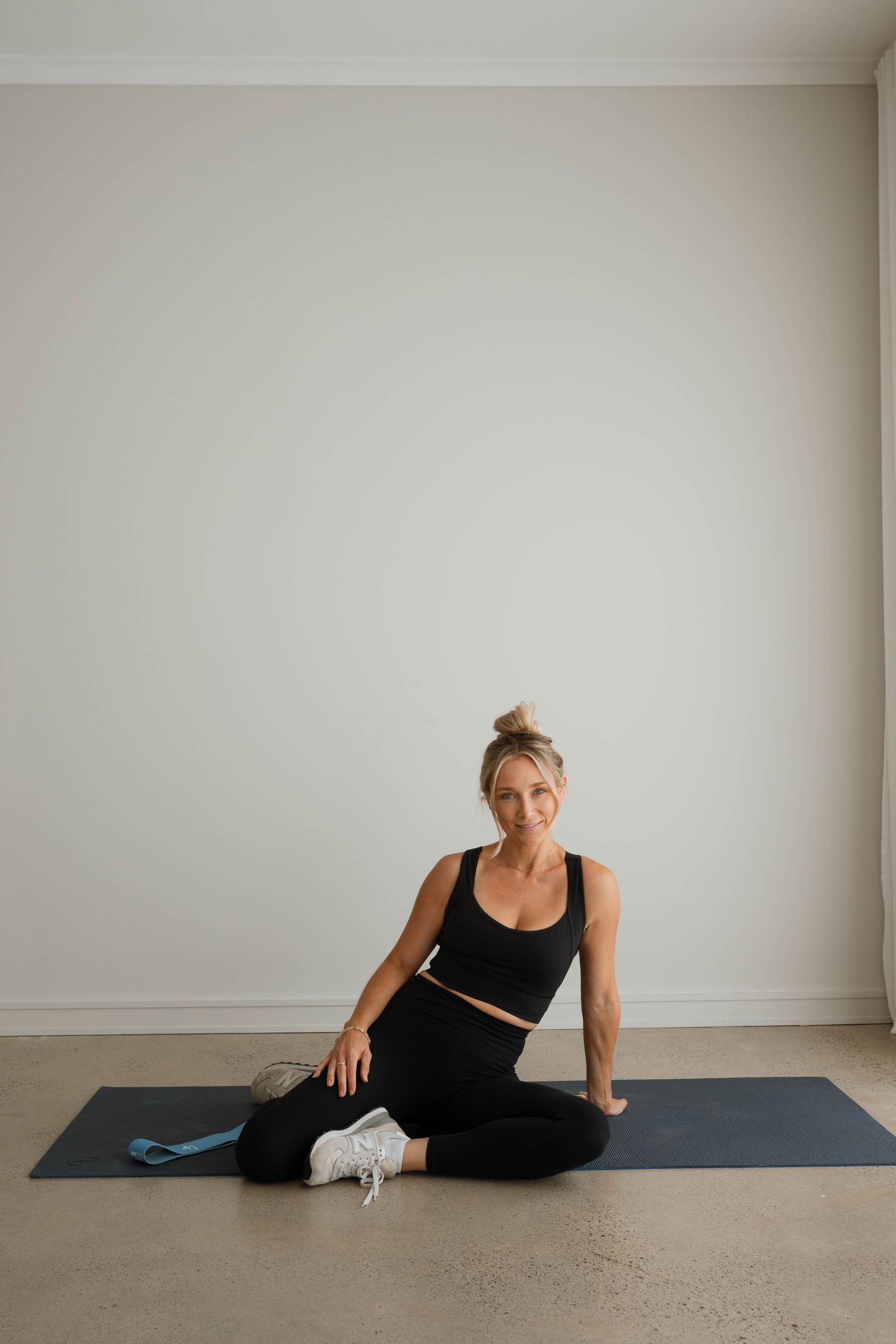 Woman in black activewear sitting cross-legged on a dark yoga mat in a minimalist white room.