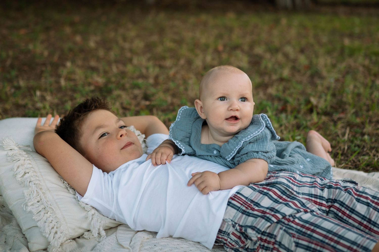 Two brothers lying on a blanket in the grass, with the older boy relaxing beside his curious baby sibling.