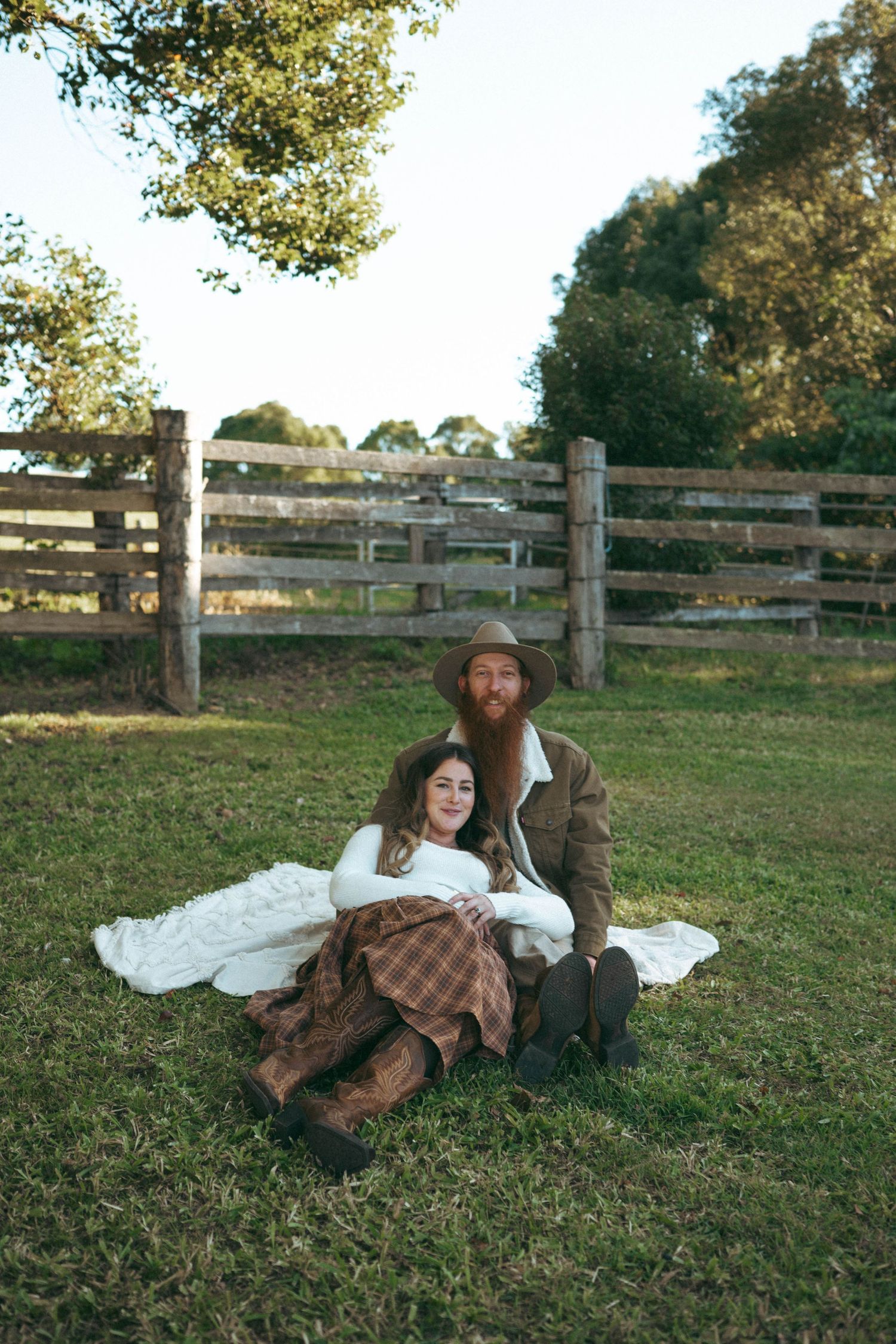 Couple sitting on grass in rustic farm setting, man with red beard in hat embracing woman in plaid skirt near wooden fence.