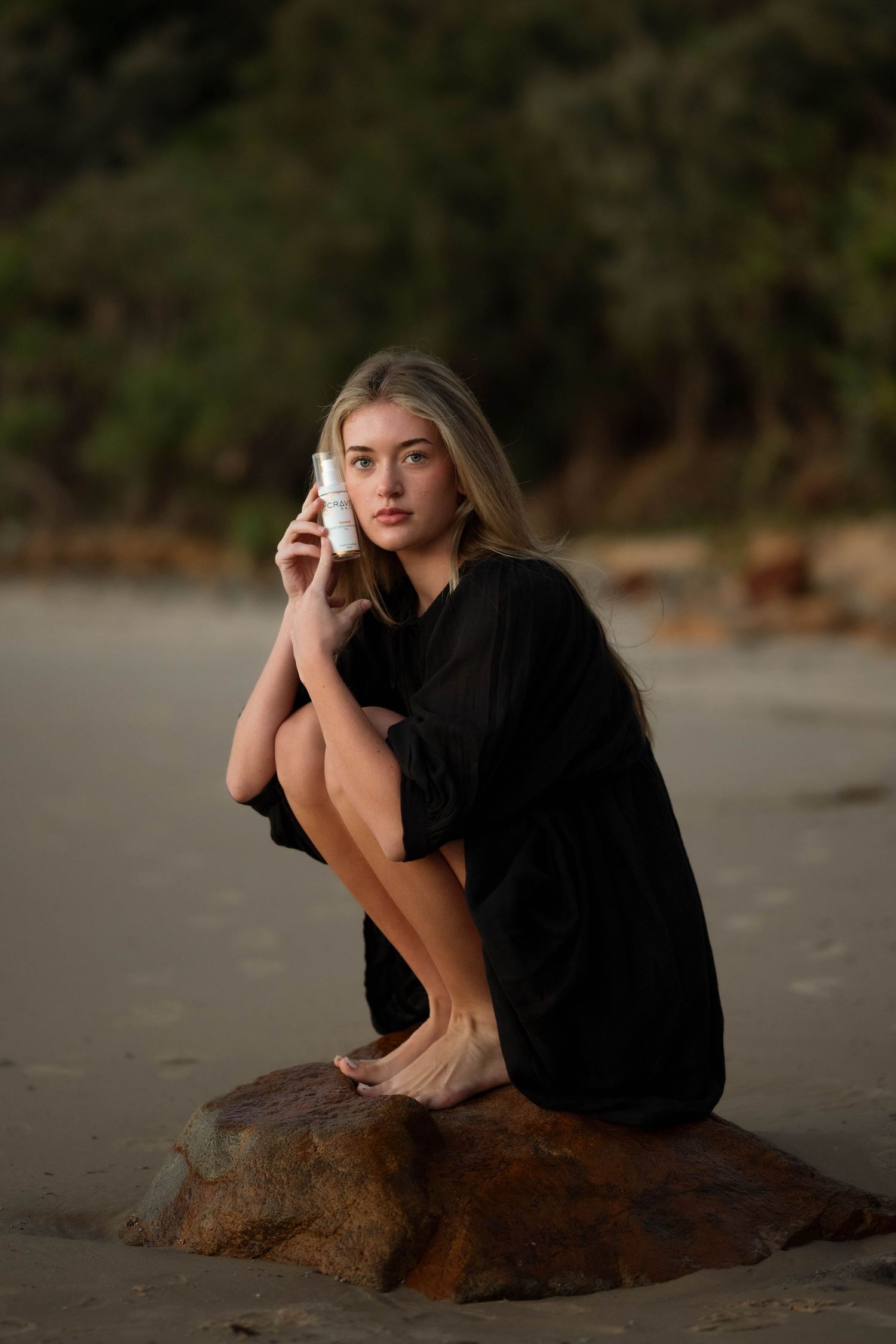 Young blonde woman in black dress sits on beach rock holding white phone, with lush green trees in background.