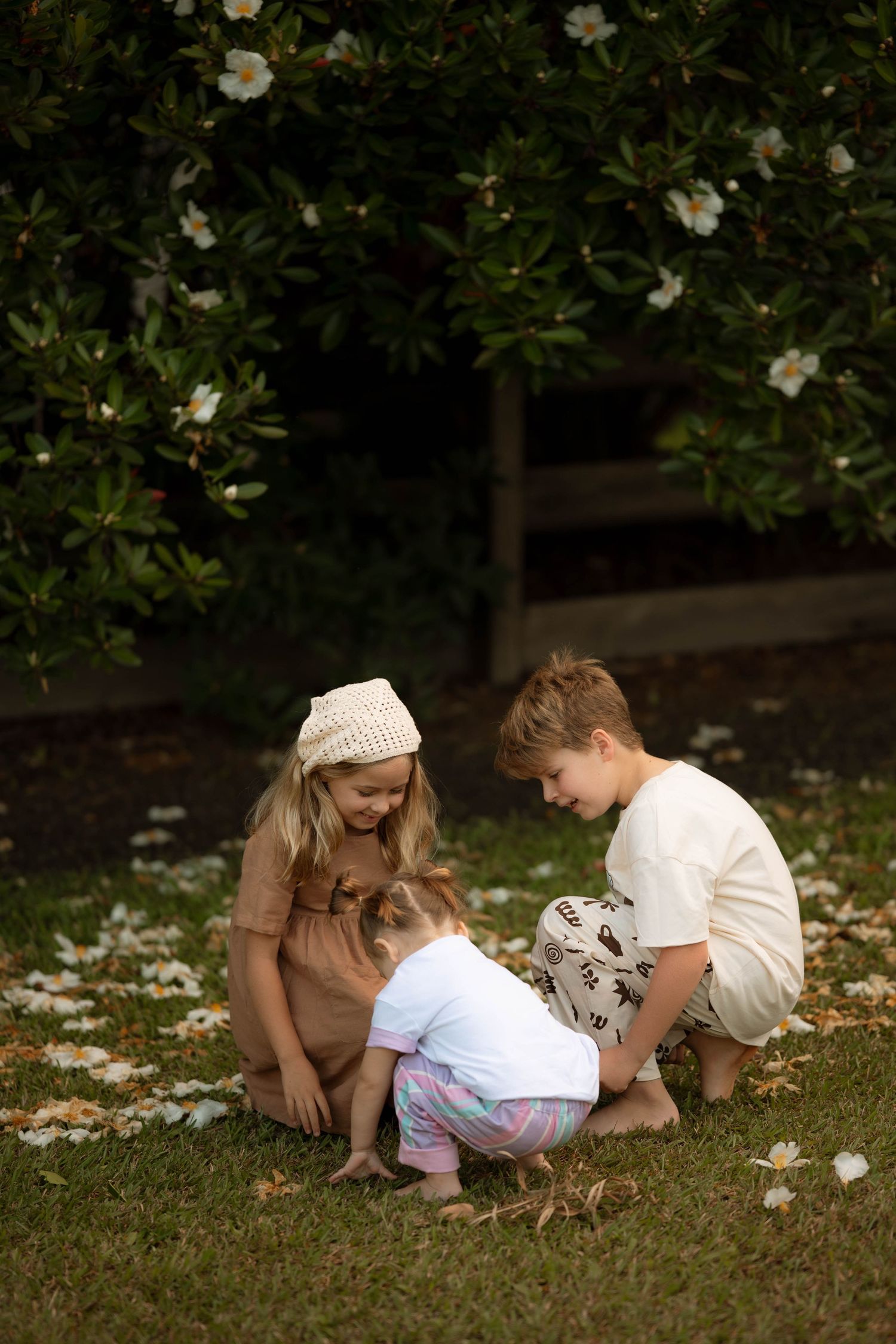 Three children playing together on grass beneath a flowering white magnolia tree outdoors.