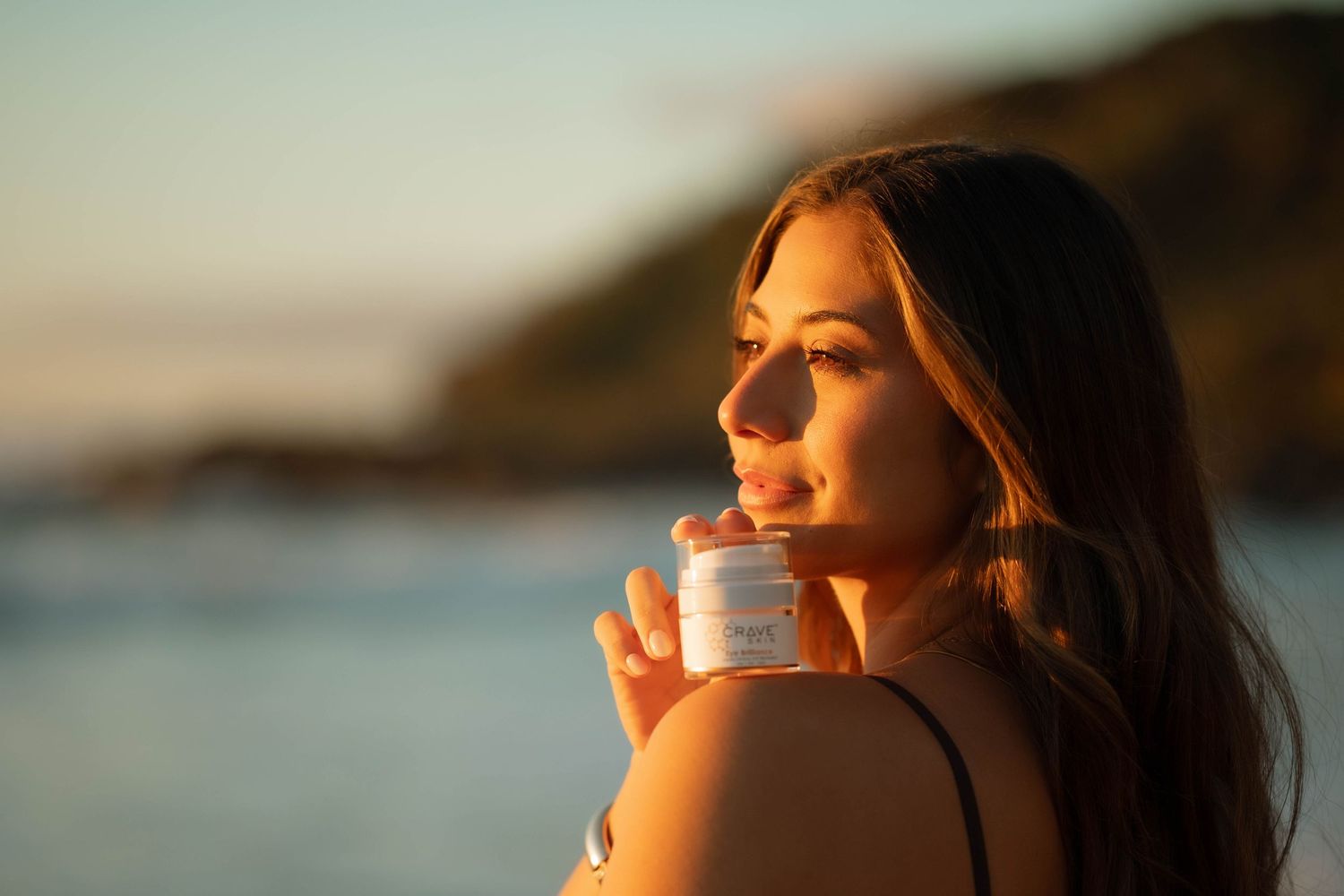 Brunette woman applying Crave skincare cream at sunset by the ocean.