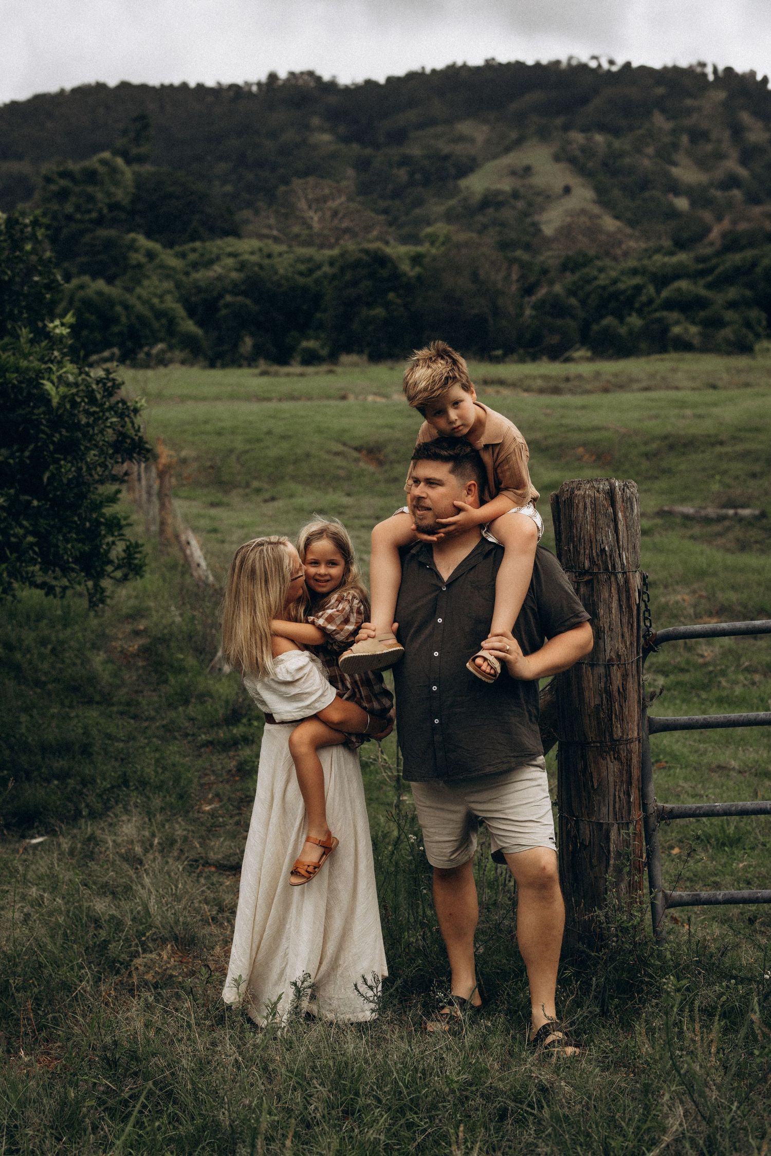 Family of four posing by rustic wooden fence in green countryside, father carrying son on shoulders.