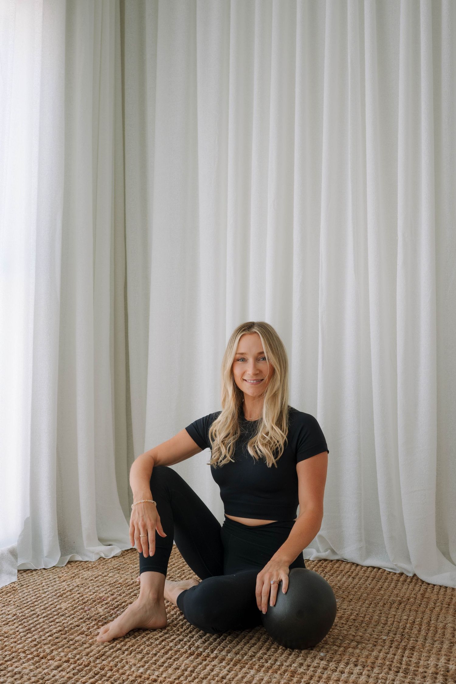 Smiling blonde woman in black crop top and leggings sitting on woven rug with pilates ball.