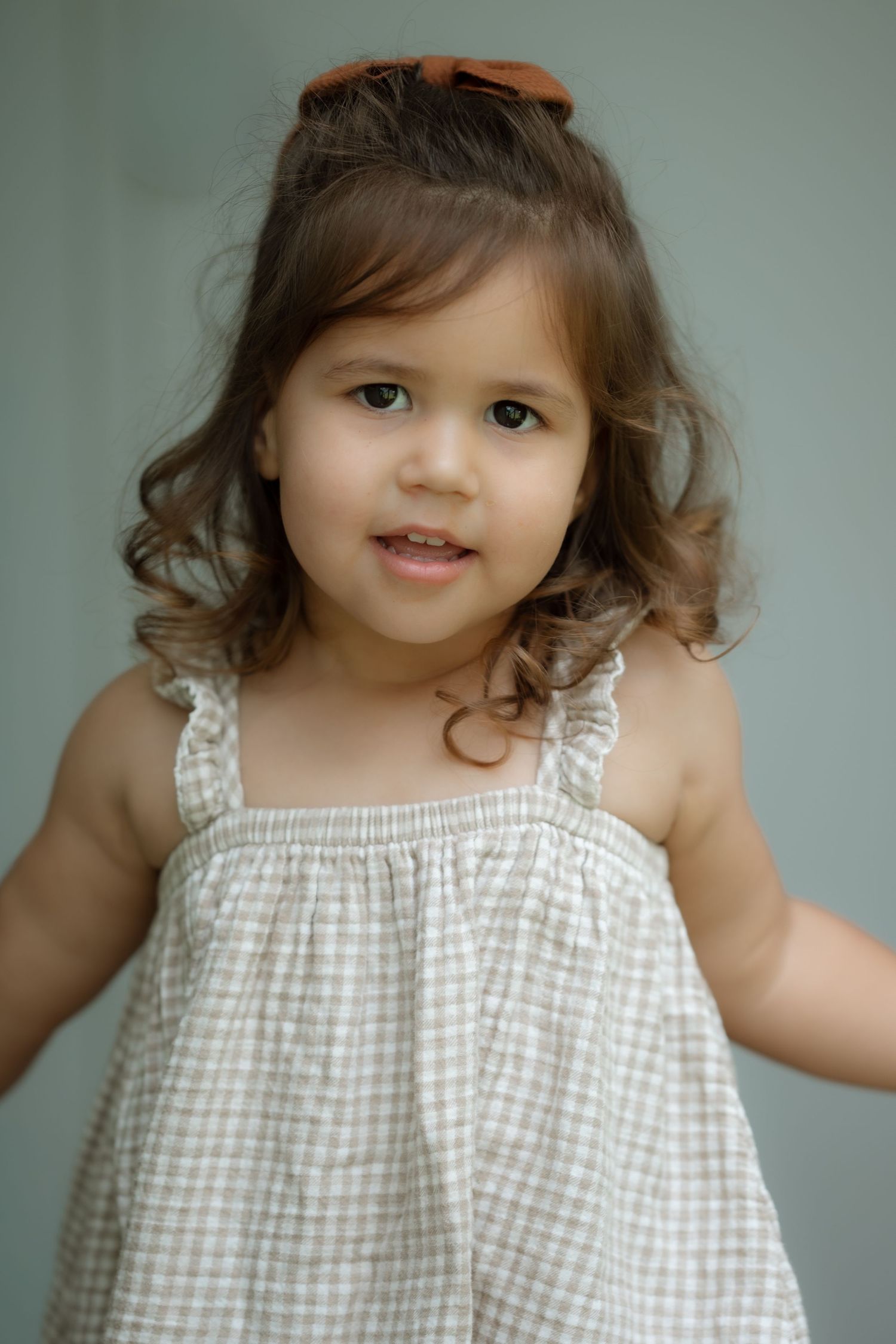 Adorable toddler girl with brown bow in hair wearing checkered sundress smiling at camera.