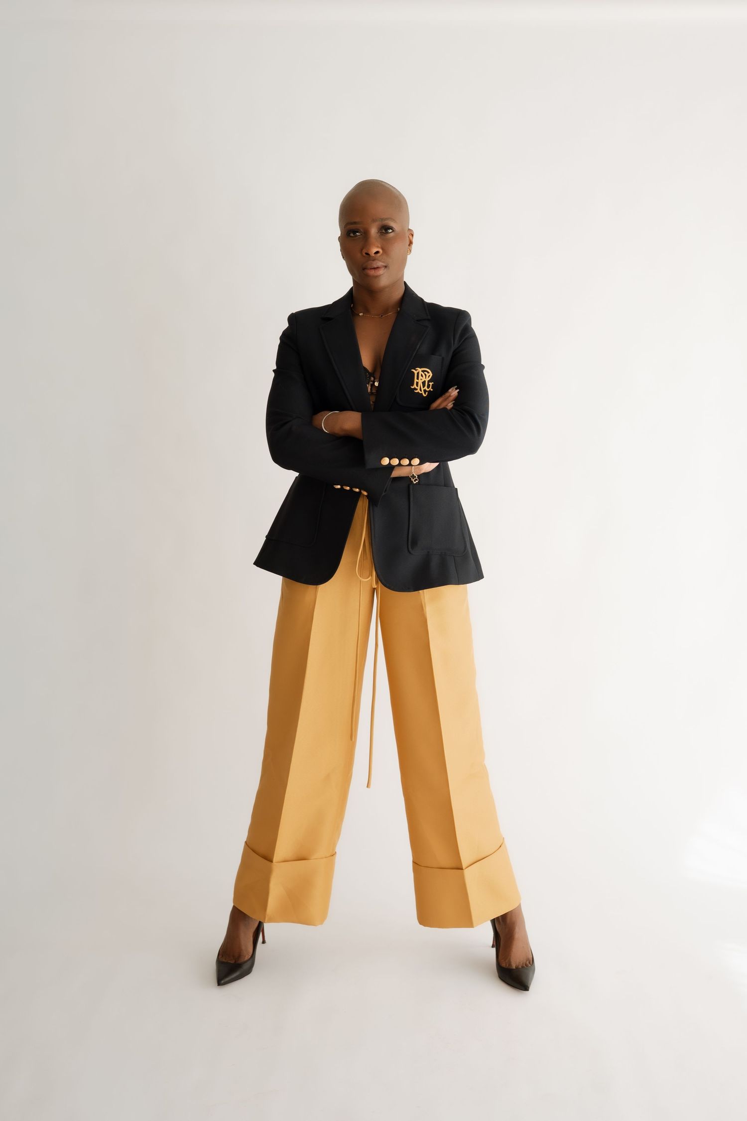 A confident bald woman poses in a black blazer with gold crest, mustard wide-leg trousers, and black heels.
