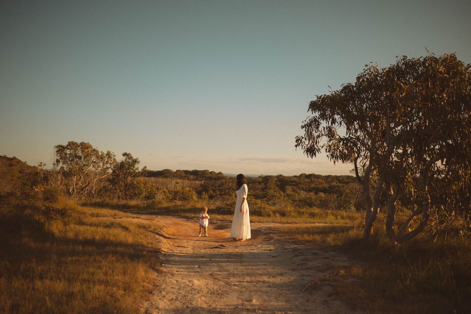 Woman in white dress walking with small dog on a dirt path at golden hour in an Australian outback landscape.