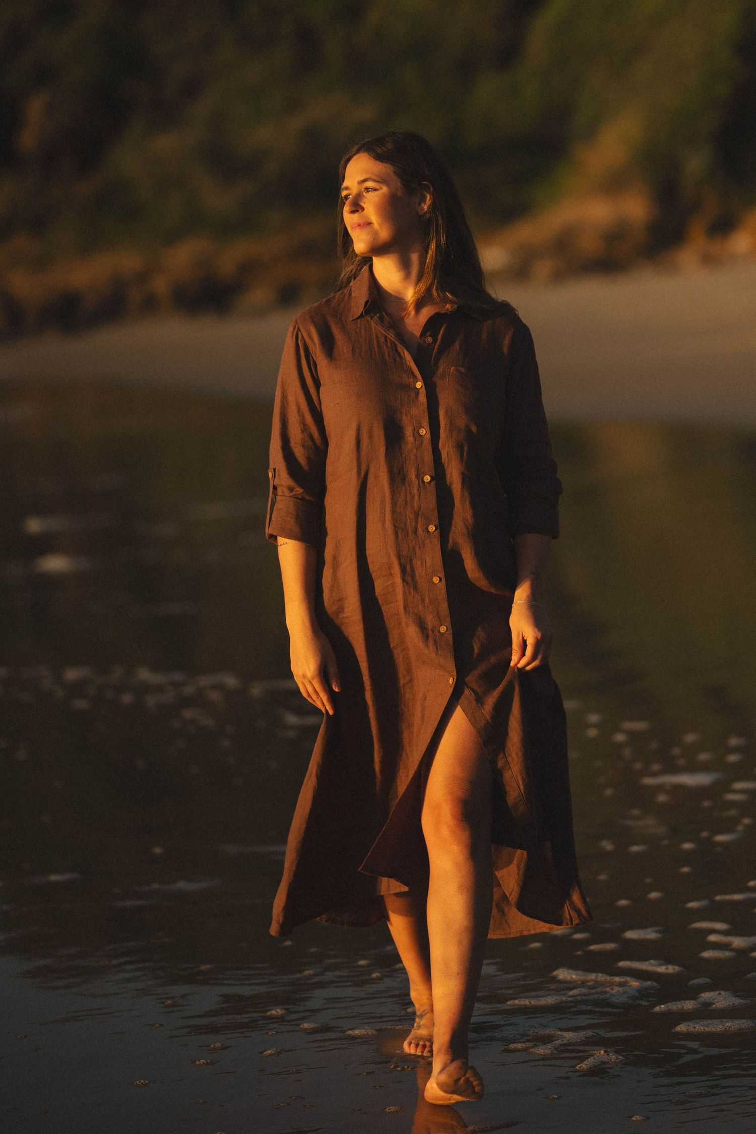 Woman in brown linen shirt dress walking on beach at golden sunset hour with green cliffs in background.