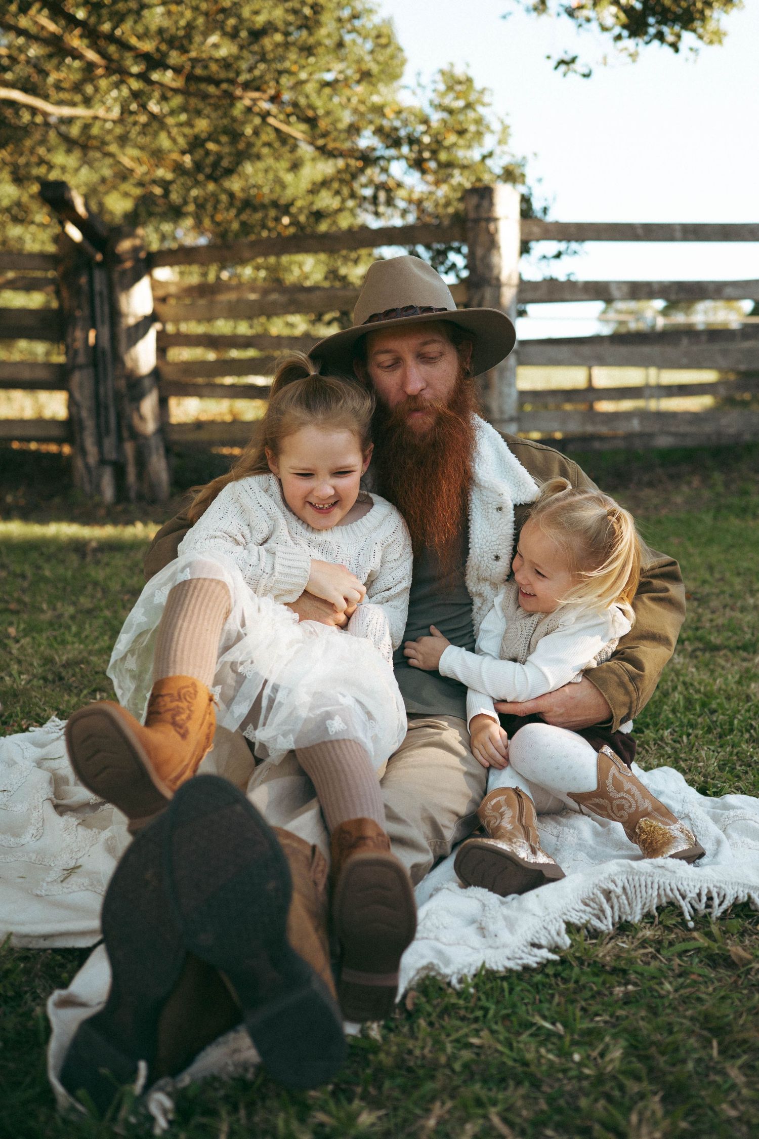 A bearded father in a cowboy hat sits on a blanket outdoors laughing with his two young daughters near a wooden fence.