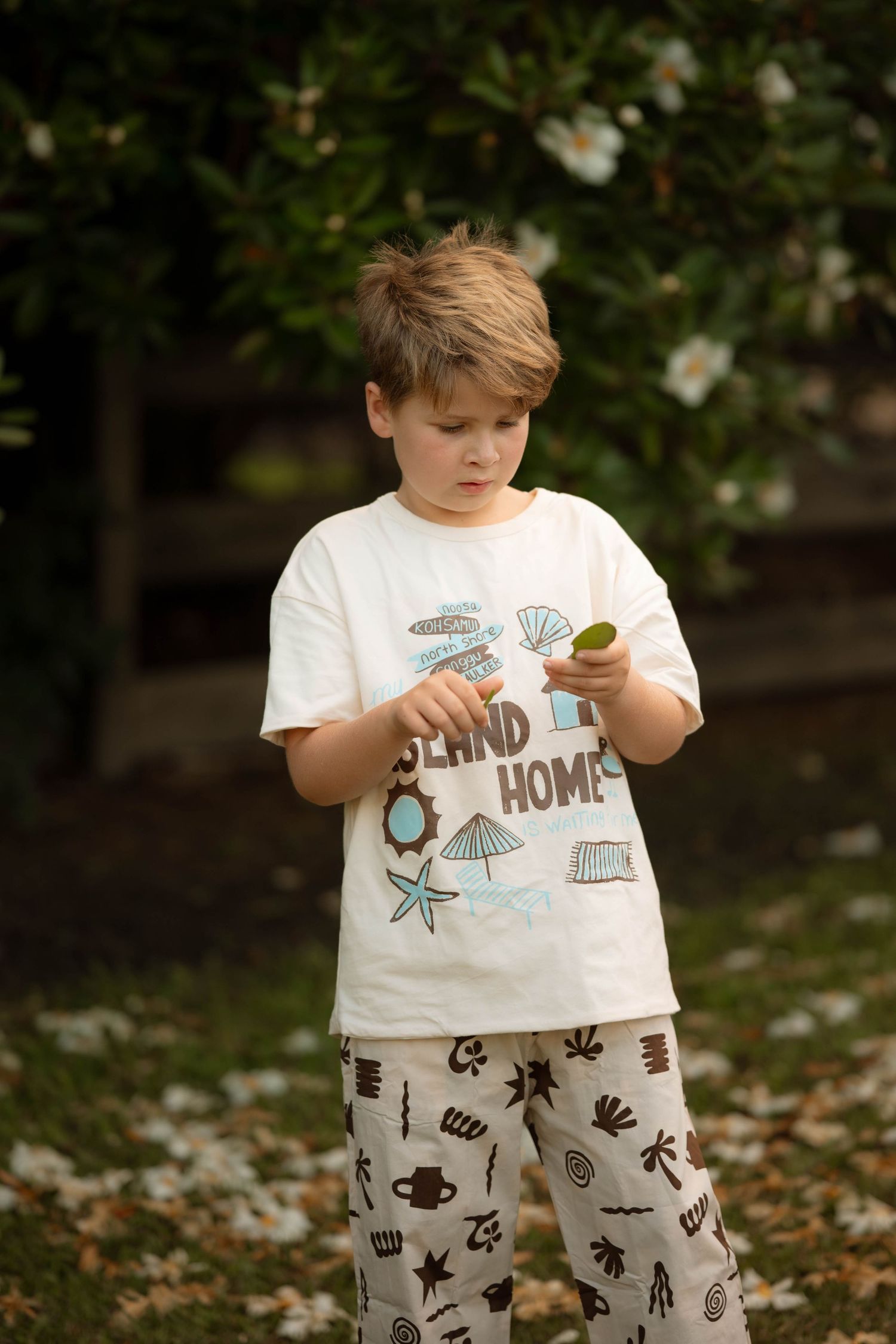 Boy in Island Home t-shirt and patterned pants examines small object outdoors near flowering shrubs.