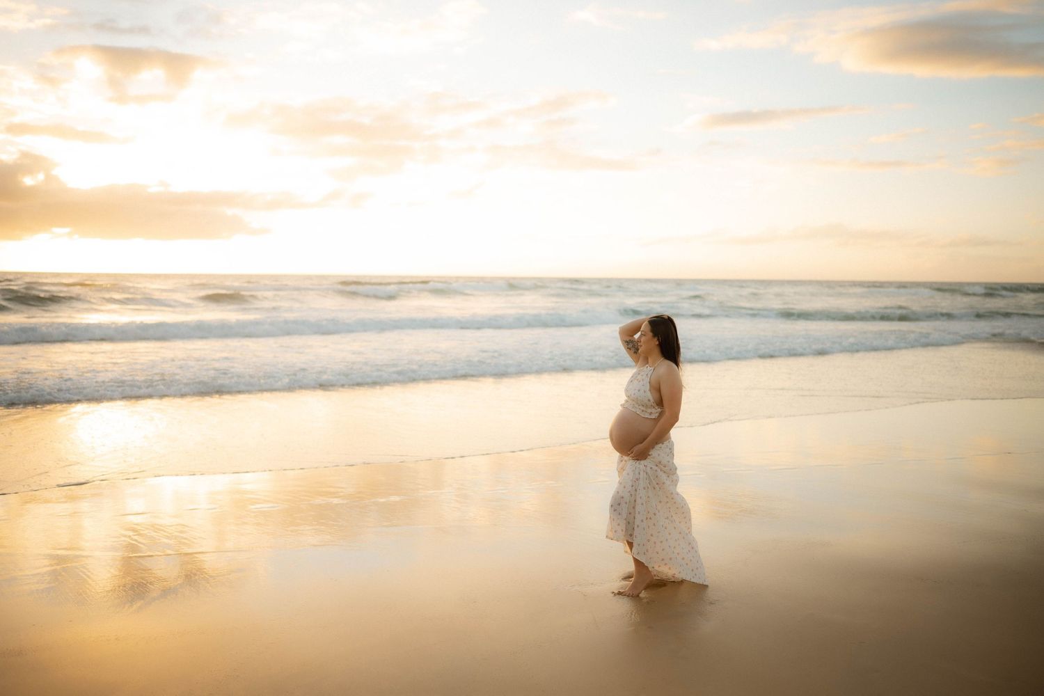 Pregnant woman in flowing white floral dress walking along sandy beach with stunning pastel sunset sky.