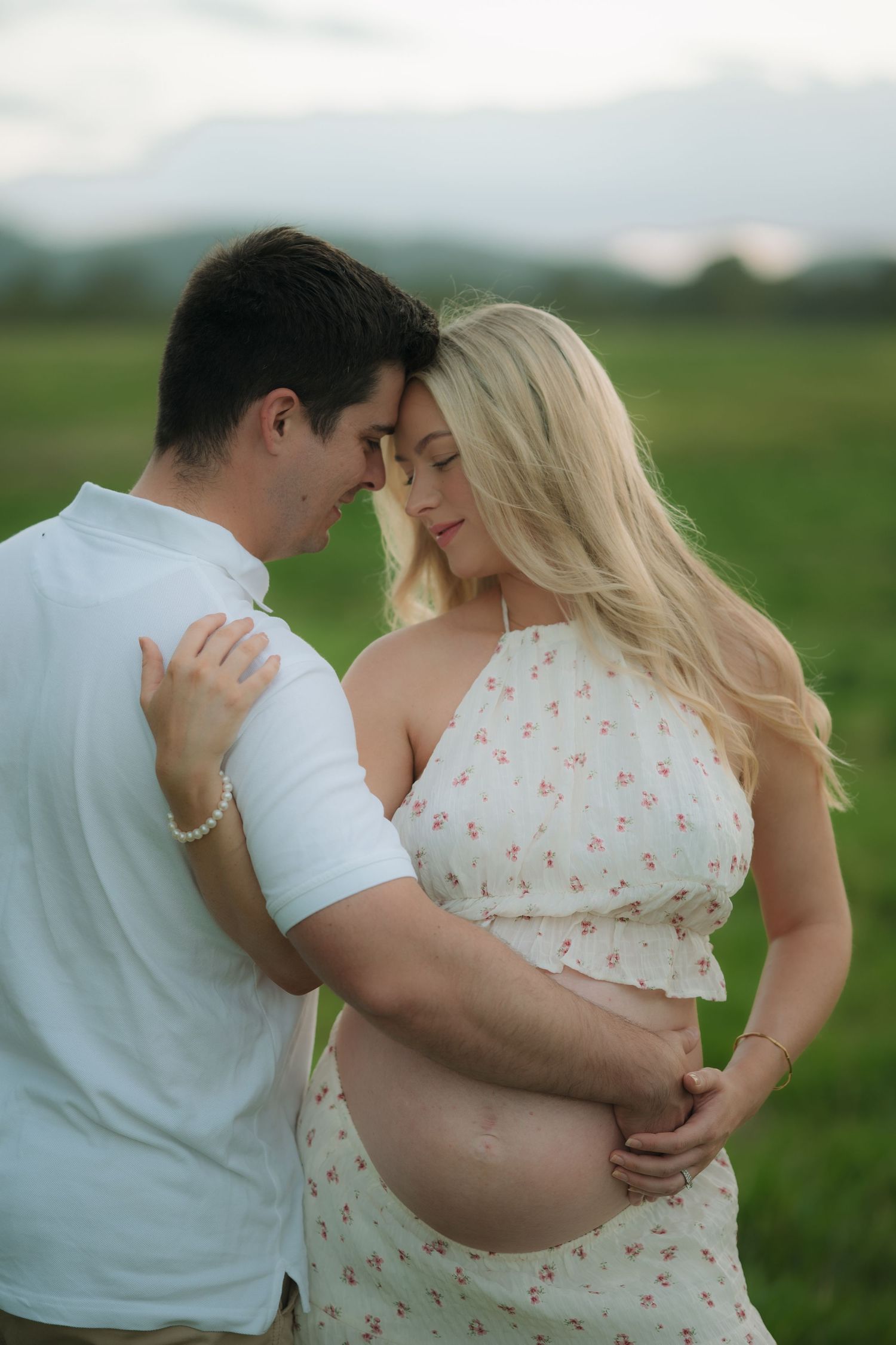 Couple sharing tender moment outdoors, man holding pregnant woman's belly in green field.