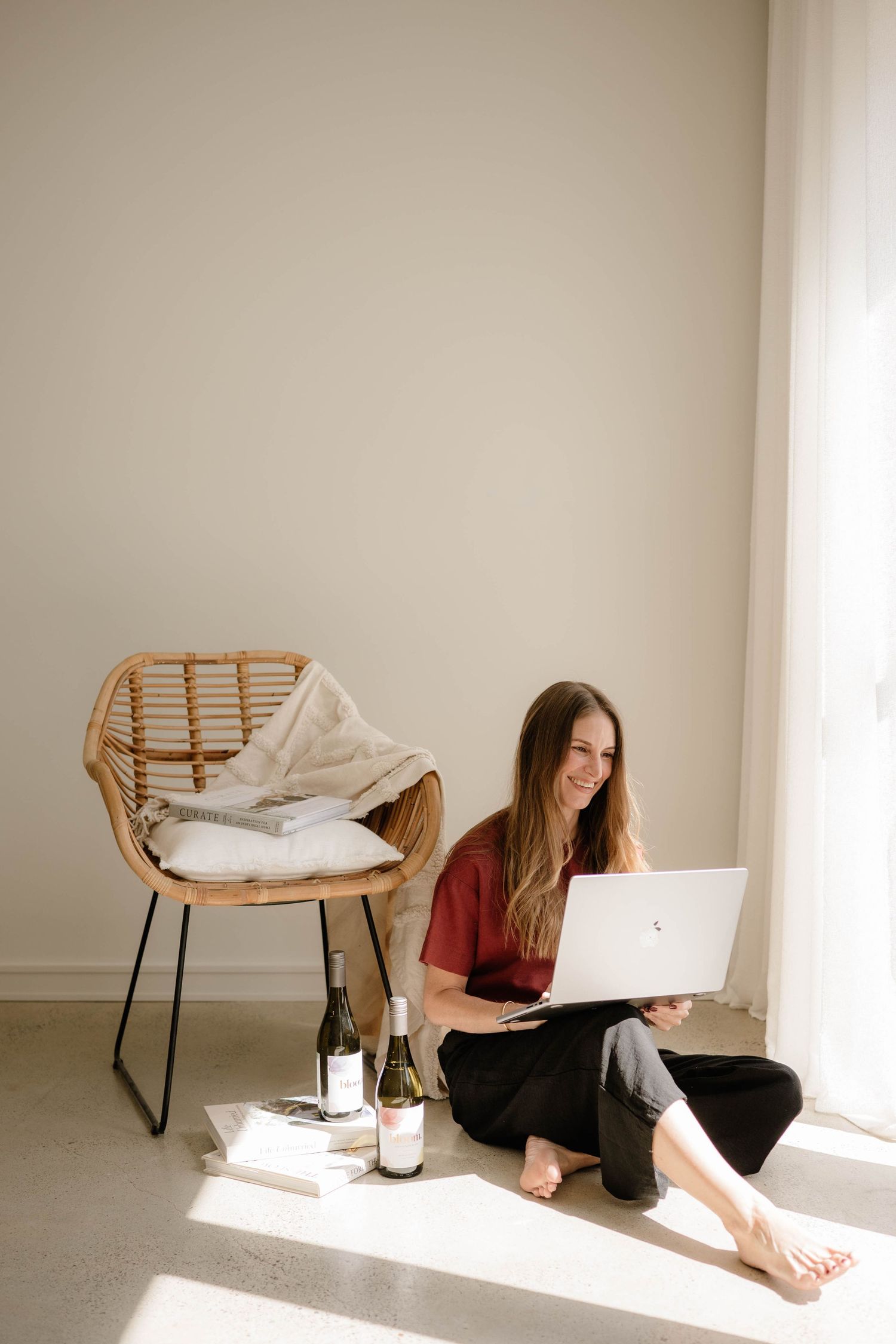 Woman in red top sitting on floor with laptop beside rattan chair and wine bottles.