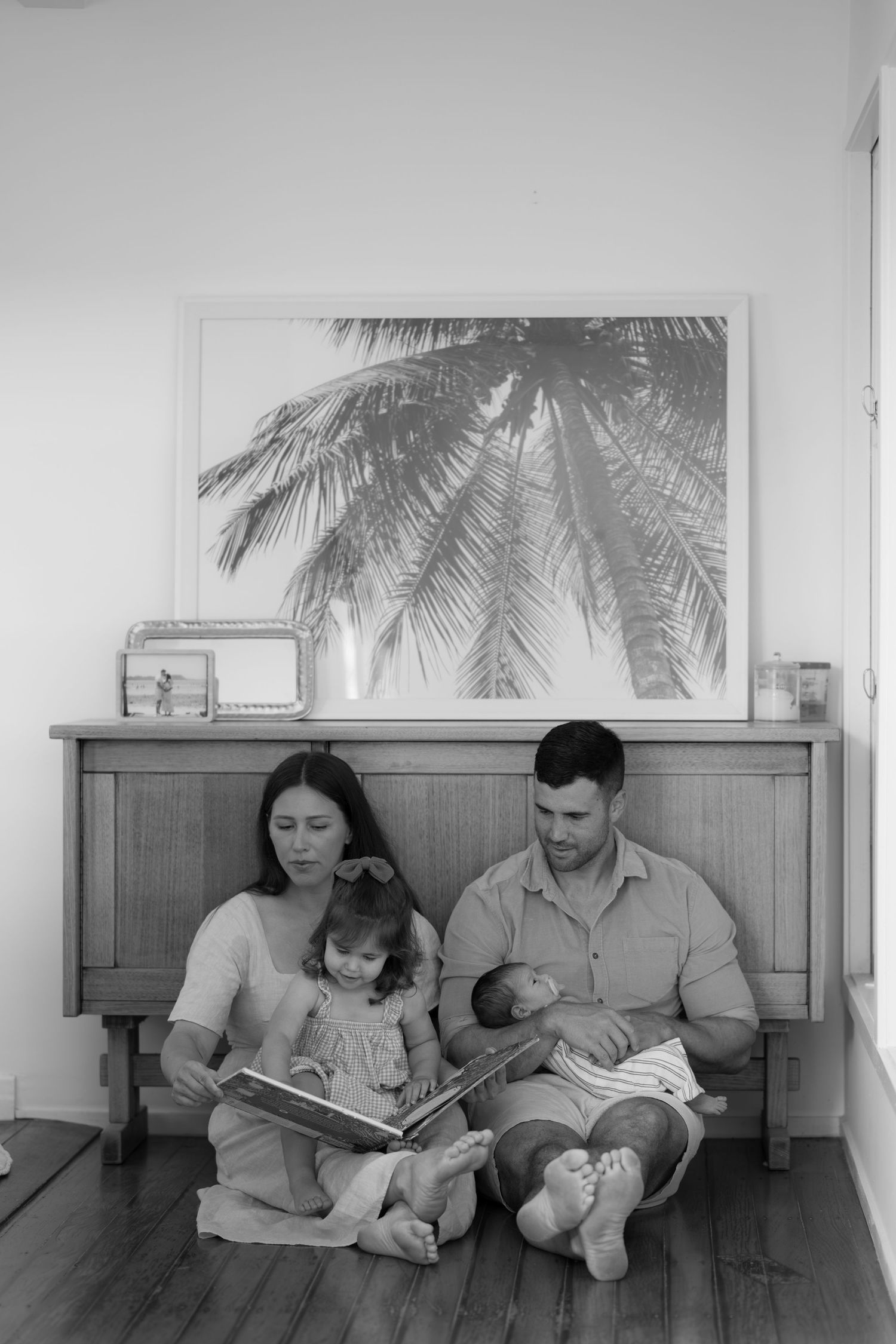 Black and white family photo with parents reading to toddler and holding newborn baby indoors.