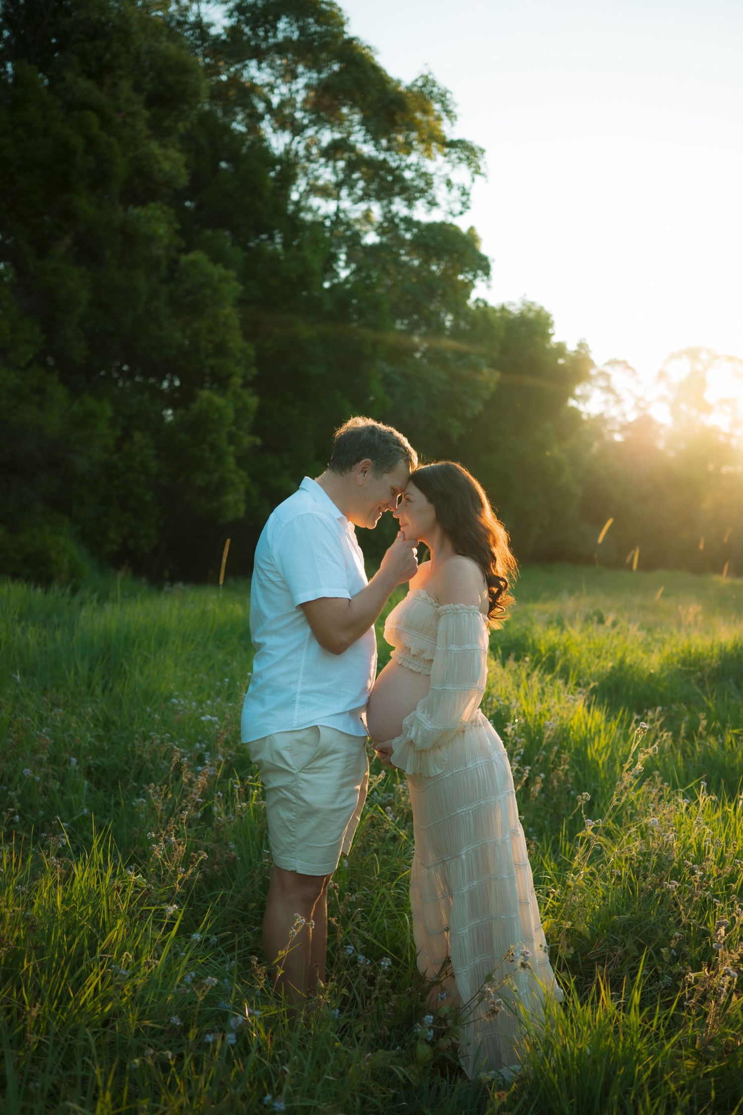 Couple sharing tender moment in sunlit green field, pregnant woman in white dress embraced by partner.