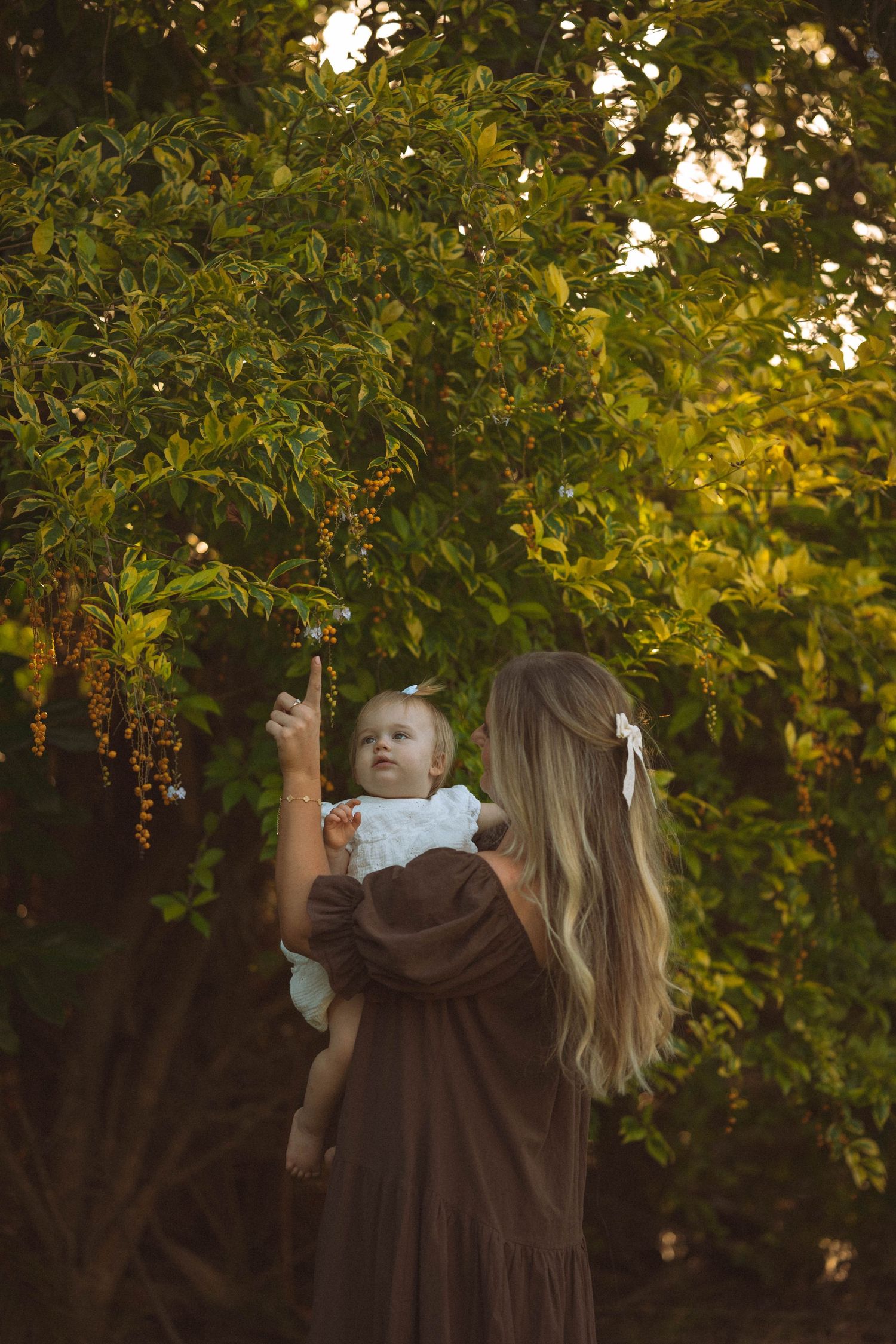 A woman with long blonde hair in a brown dress holds her baby up toward green tree branches at golden hour.