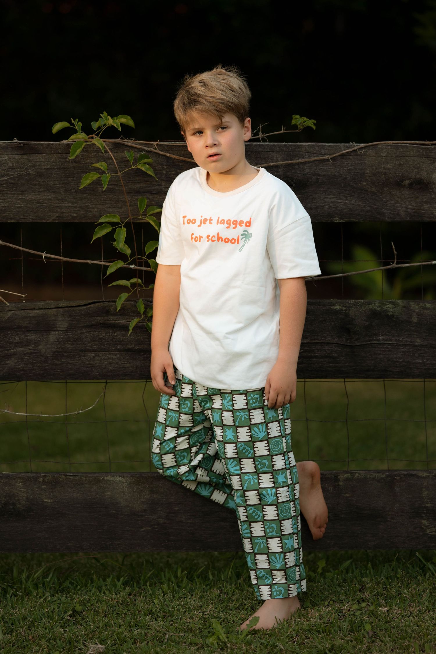 Young boy in white graphic tee and patterned jogger pants leaning against a wooden fence outdoors.