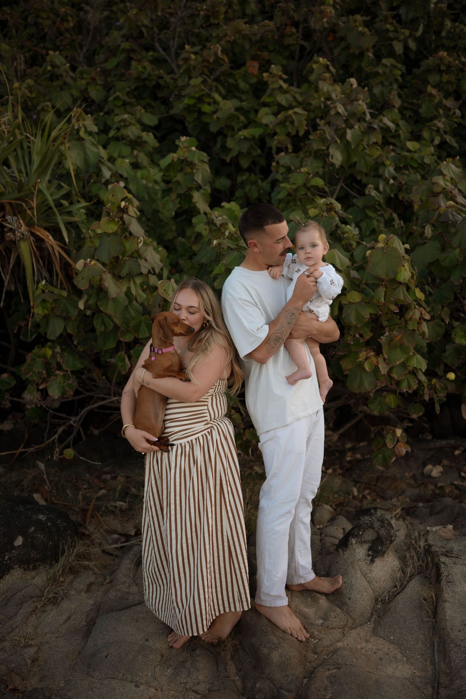 A young family poses outdoors, woman in striped dress holds a dog, man in white holds a baby, with lush greenery behind them.