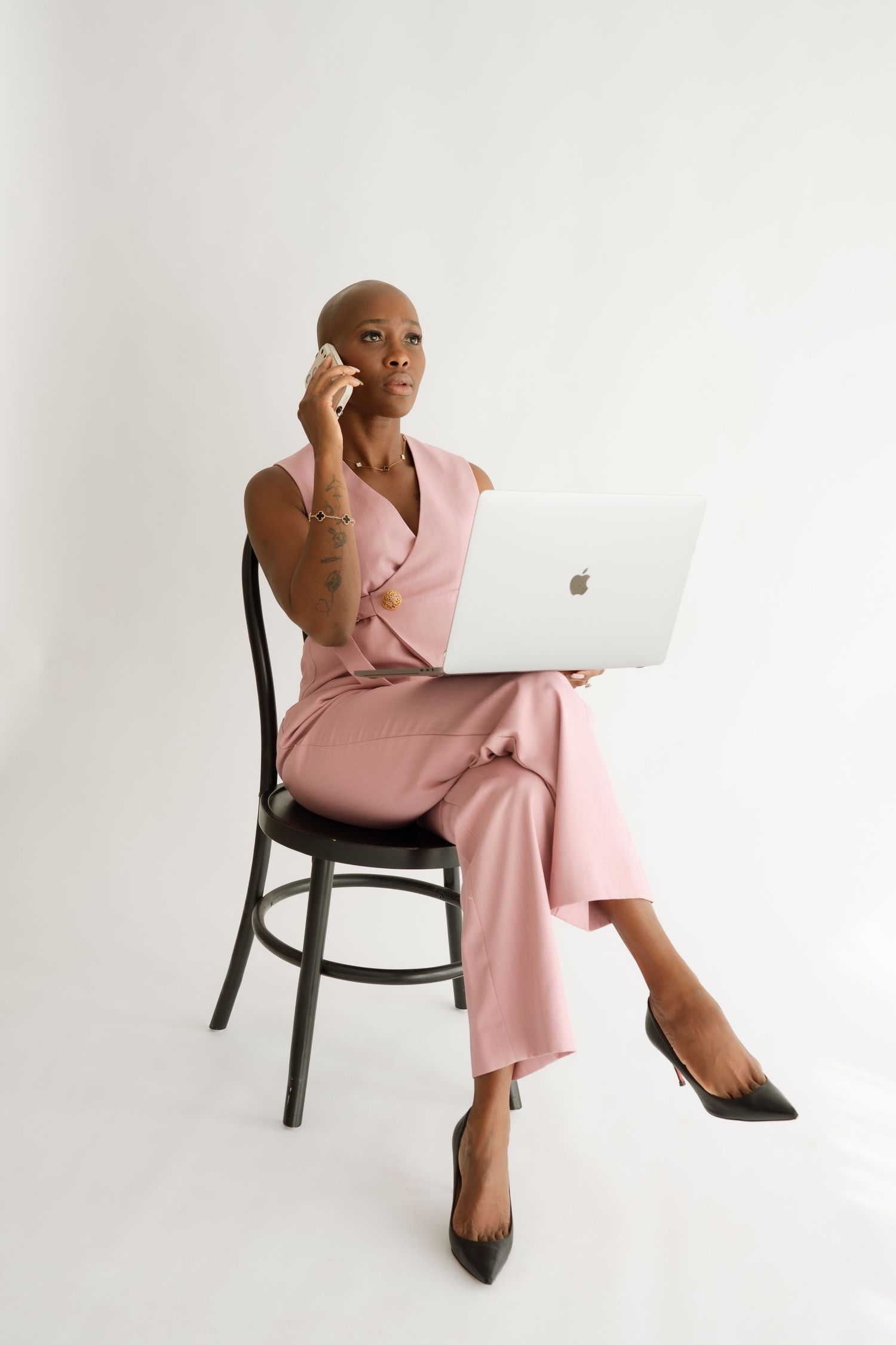 A bald Black woman in a pink pantsuit sits on a chair, talking on phone while using a MacBook laptop.