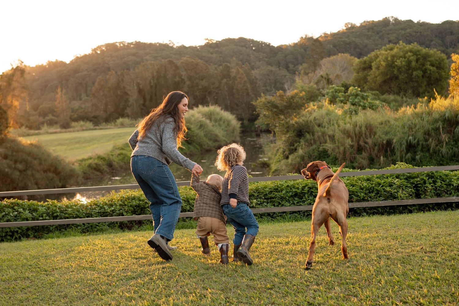 Woman and child playing with a brown dog on a green lawn at golden hour with lush hills in the background.