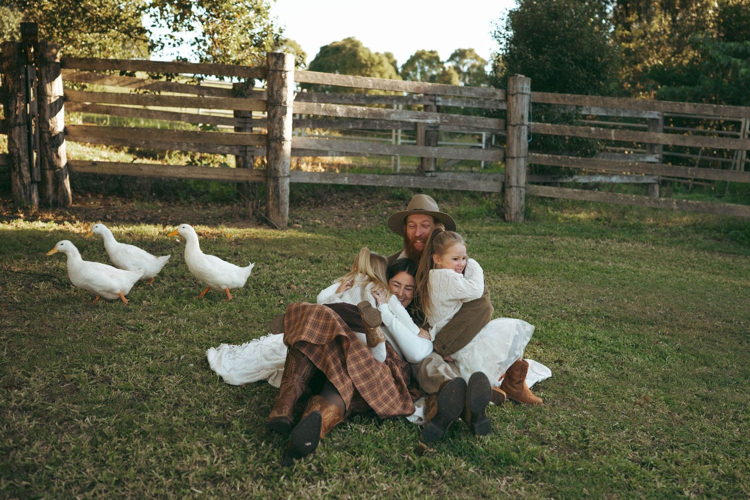 Family sitting together on grass near white geese and wooden fence on a sunny farm day.