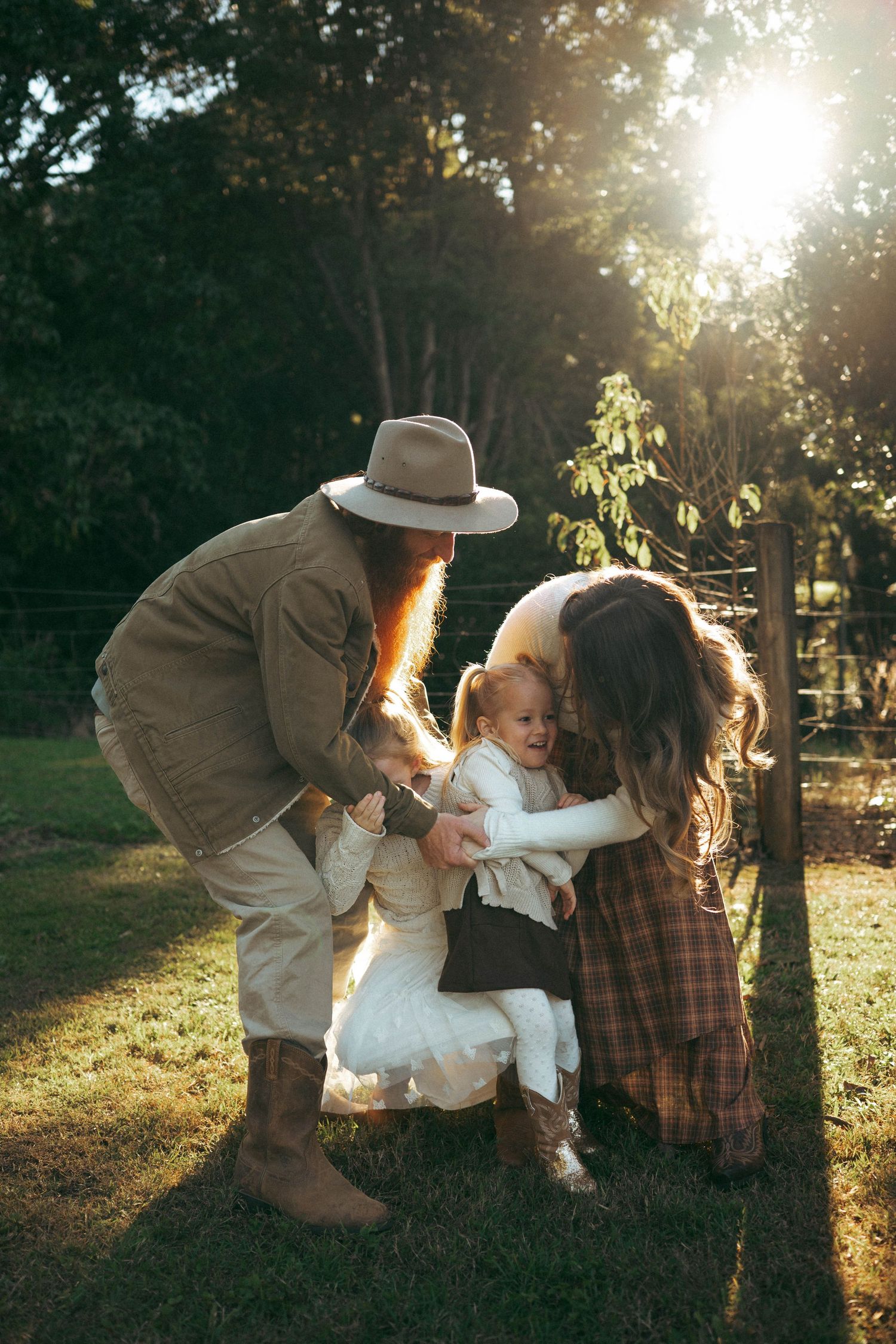 Family with red-bearded father in hat and mother bonding with baby outdoors in golden sunlight on green grass.