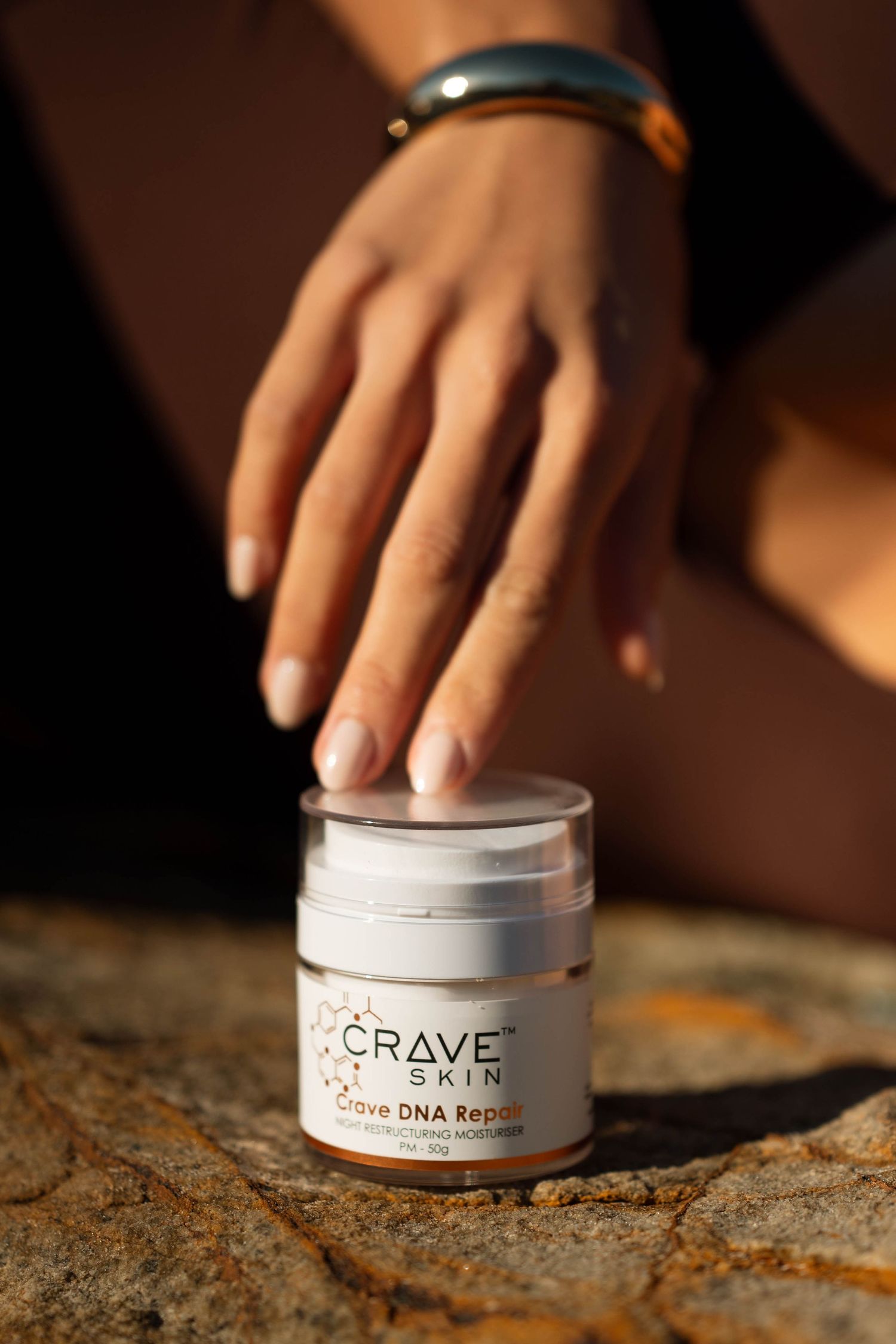 Hand reaching toward Crave Skin DNA Repair moisturizer jar on rocky surface in warm sunlight.