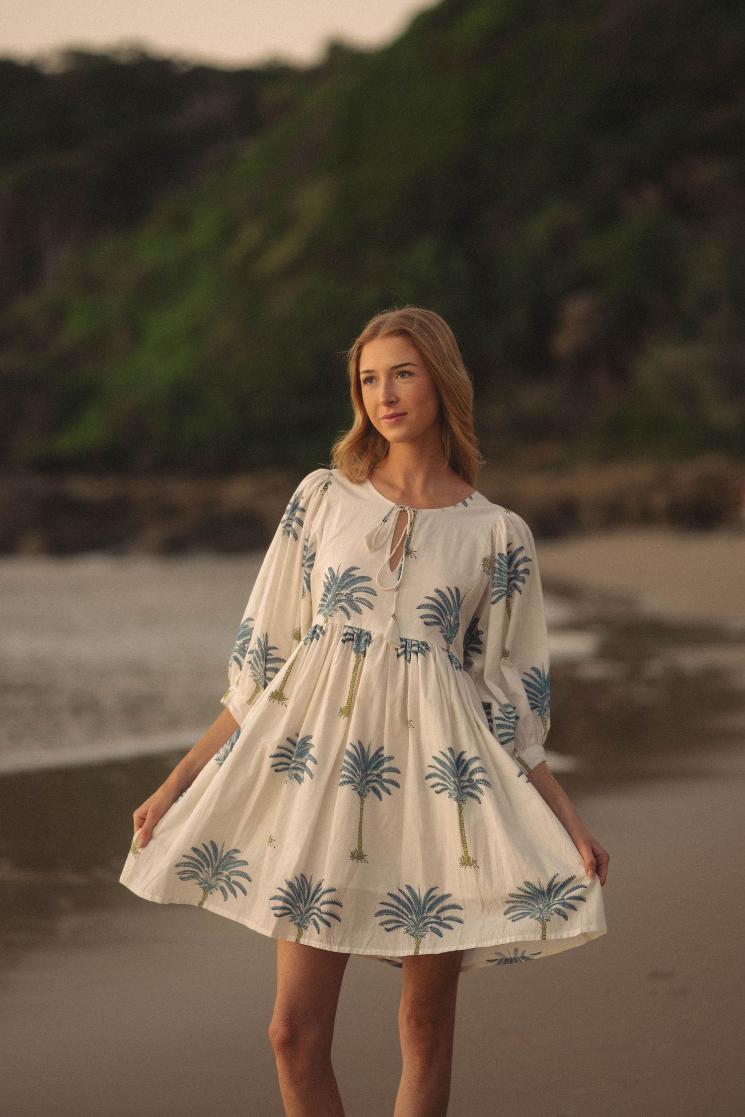 Woman in white palm tree print mini dress posing on a beach with lush green hills in the background.