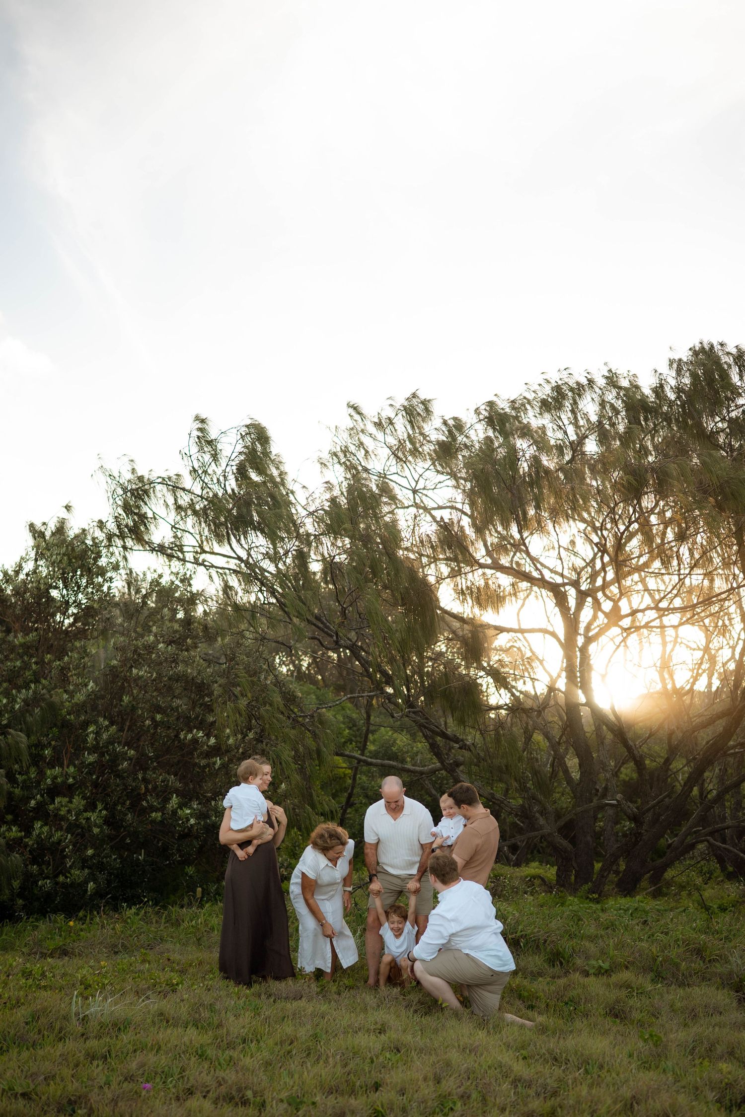 Family group gathered on a grassy field at sunset, with adults crouching down to interact with a small child.