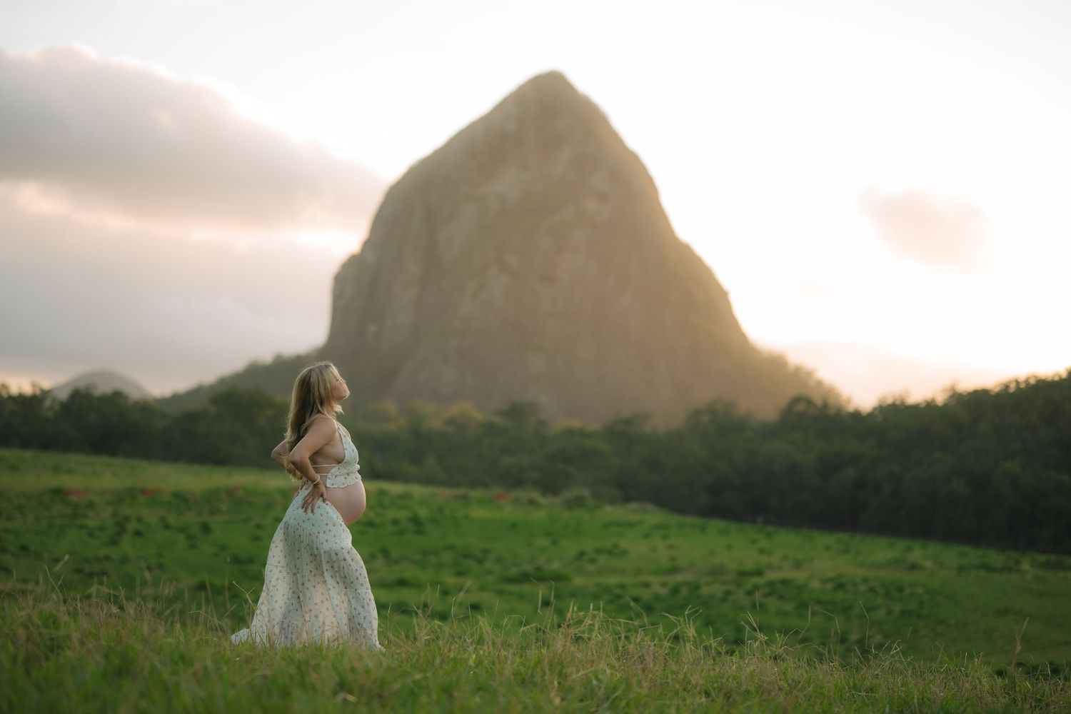 Pregnant woman in white lace gown stands in green field at sunset with dramatic rock formation behind her.