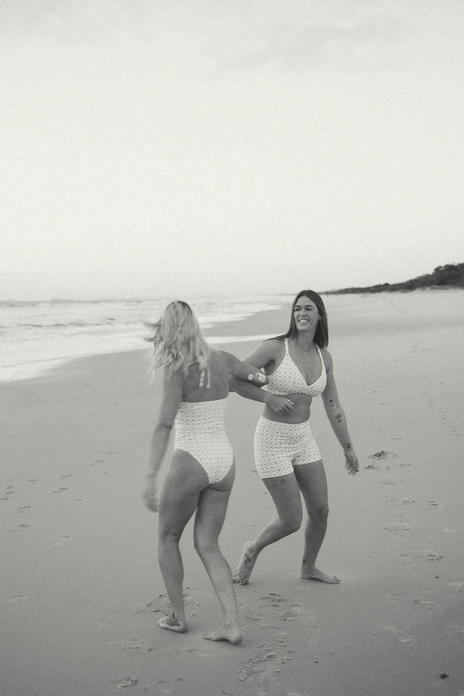 Black and white photo of two women laughing and dancing on a beach at dusk.