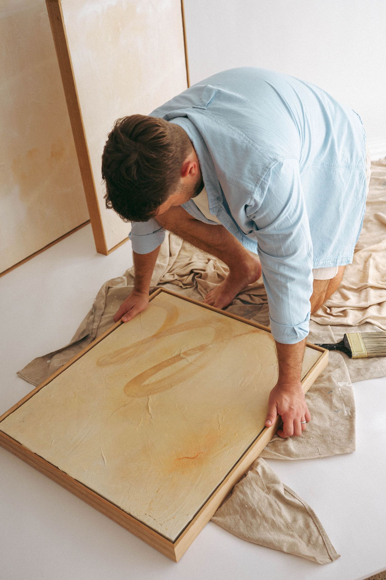 Man in blue shirt bending over a large canvas painting on the floor in a bright white art studio.