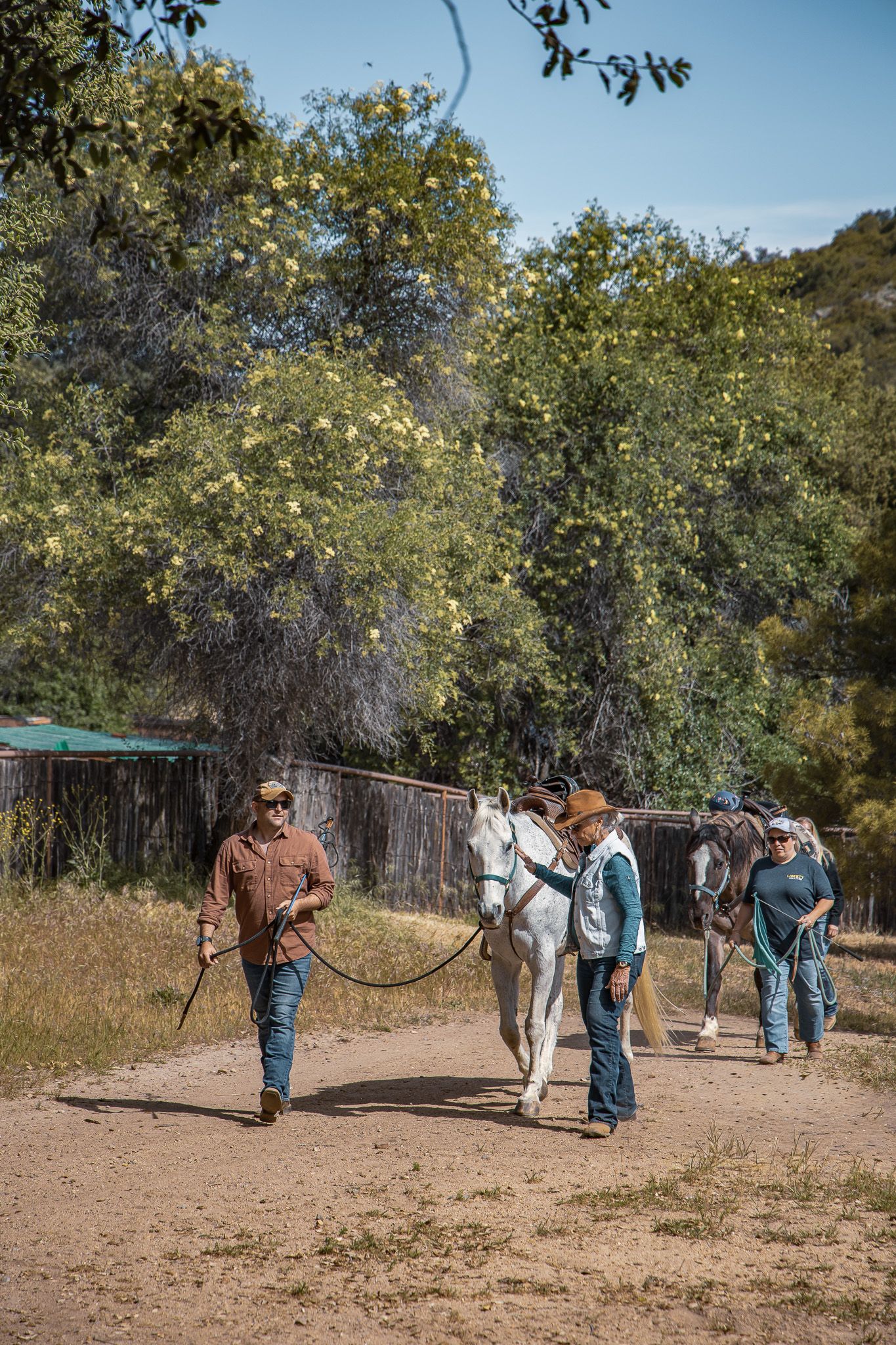 Two cowboys leading white horses along a dirt path surrounded by green trees on a sunny day.