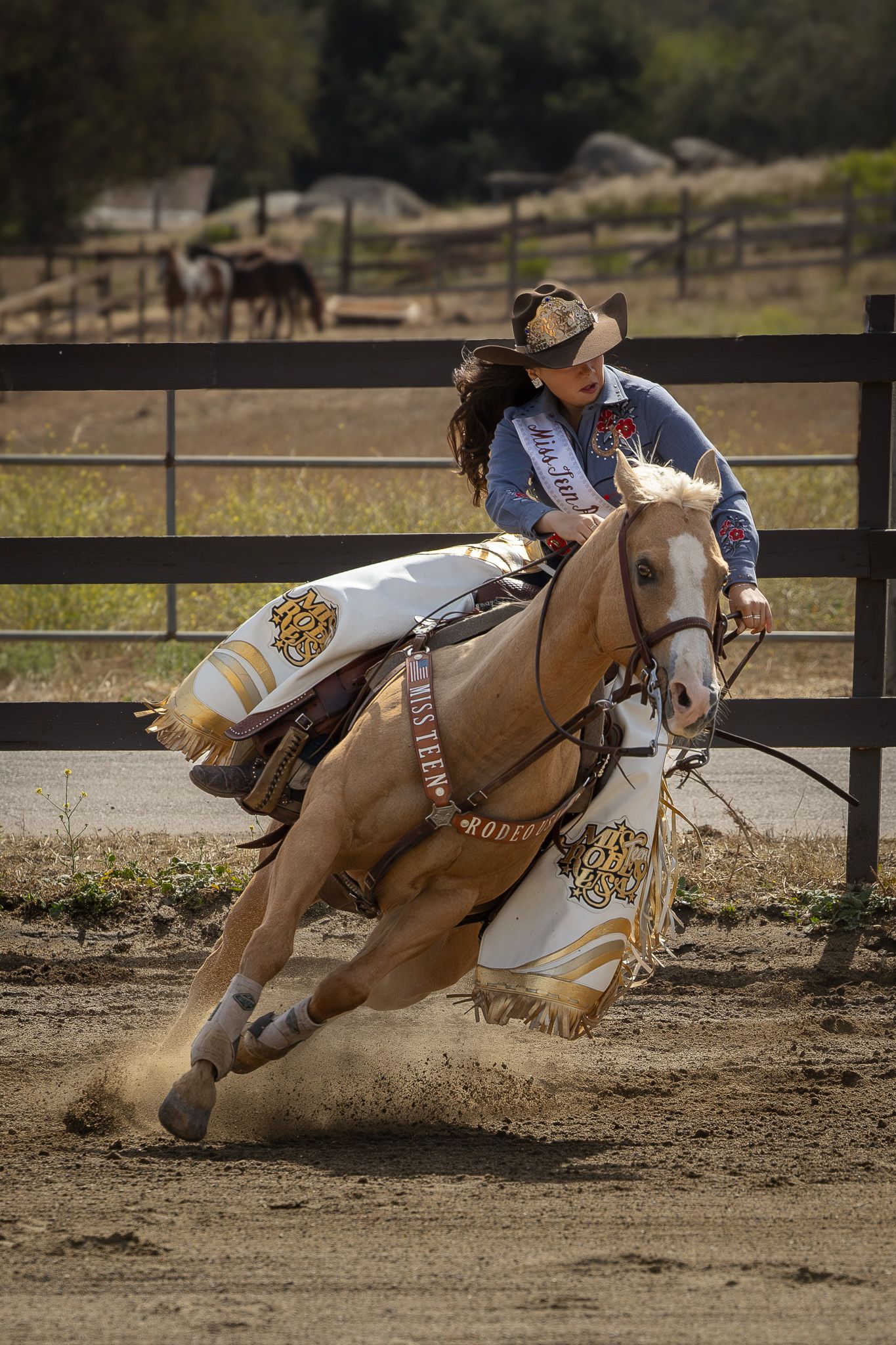 Cowgirl riding a palomino horse at full gallop during a barrel racing competition in a dirt arena.